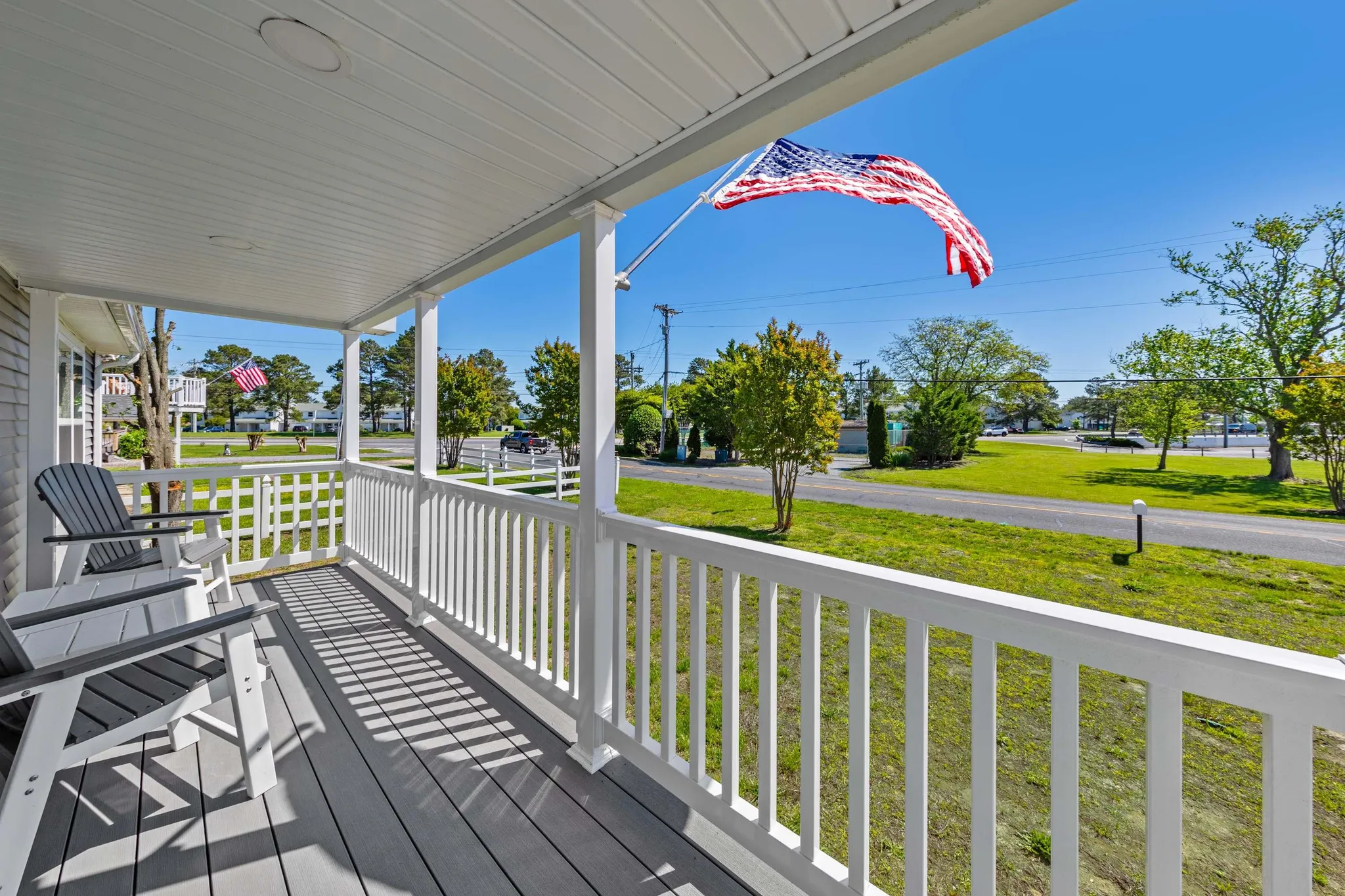 Covered porch with blue swing bench, teal Adirondack chair, and covered grill overlooking neighborhood homes under blue sky