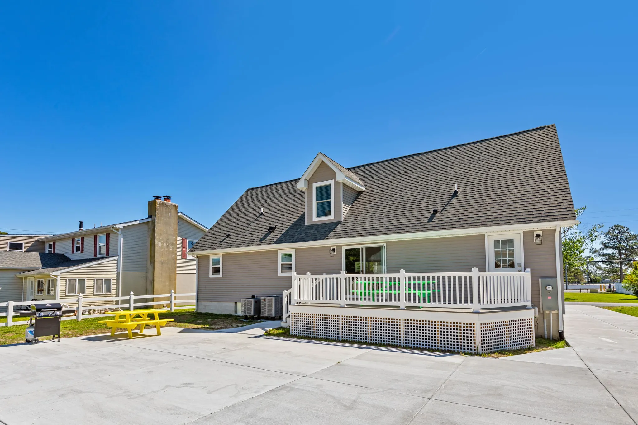 Gray two-story house with dormers and white deck railing, yellow picnic table and grill on concrete patio under blue sky