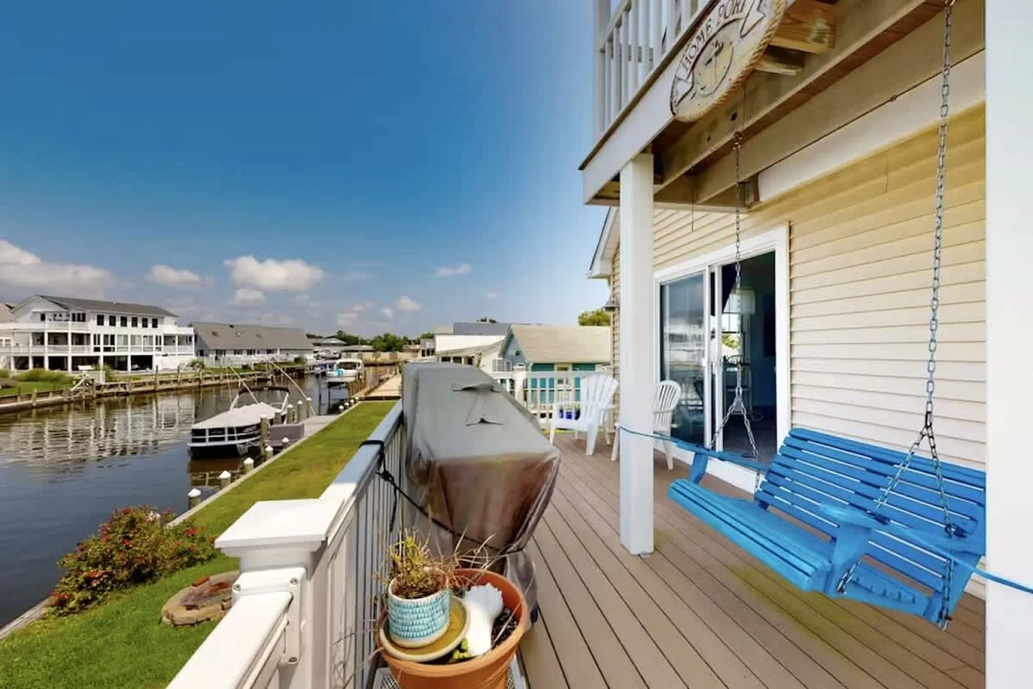 Waterfront deck with blue porch swing, covered grill, and potted plants overlooking a canal with boats and homes under blue sky