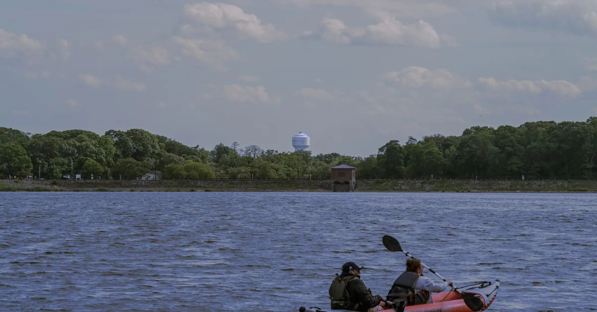 Two people paddling a kayak on a calm lake with a wooded shoreline, water tower and small building in the distance