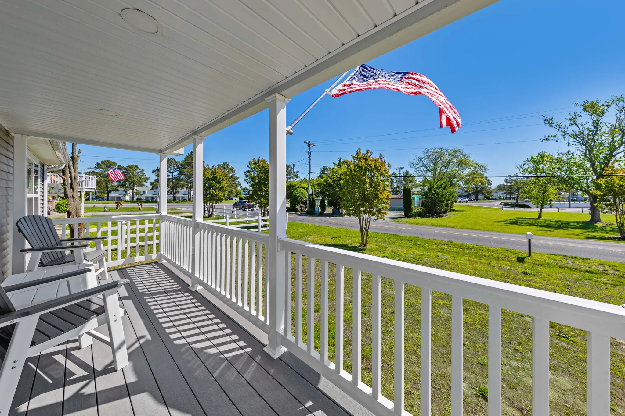 Covered porch with white railing, Adirondack chairs, and American flag overlooking a green lawn and neighborhood street