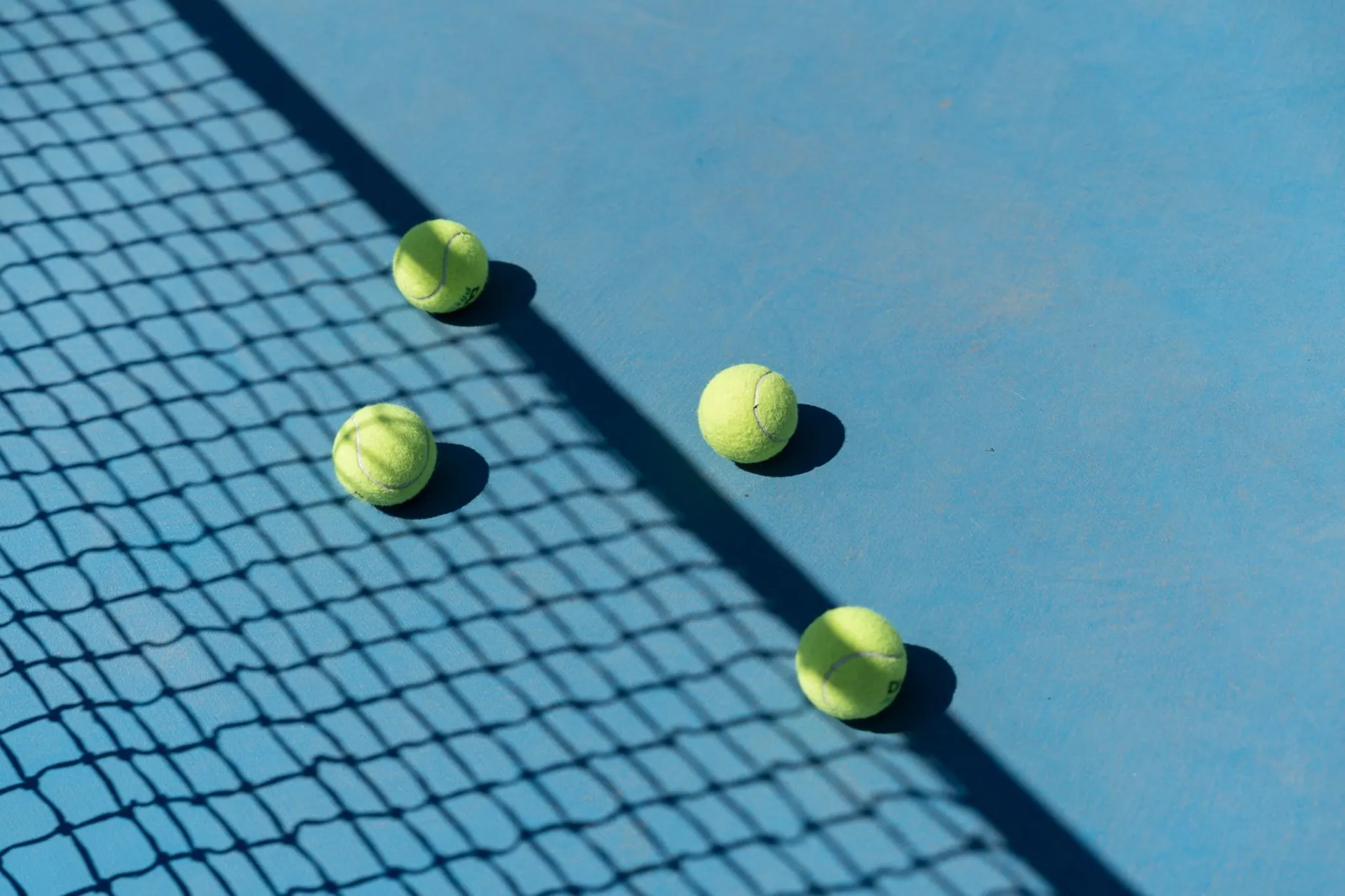Tennis balls and a net shadow on a blue hard court in bright sun