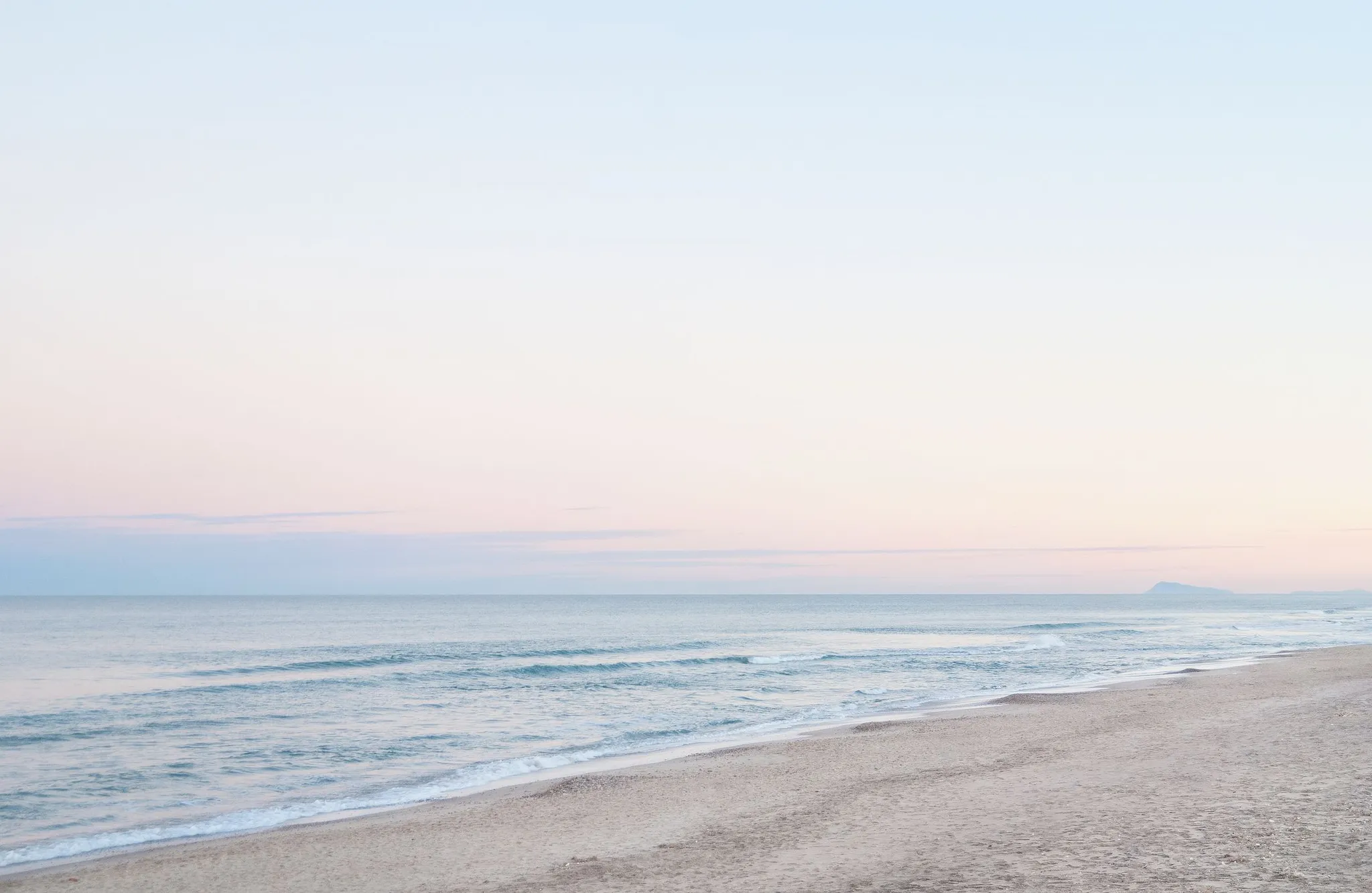 Calm sea meeting an empty sand beach under a soft pastel dawn sky