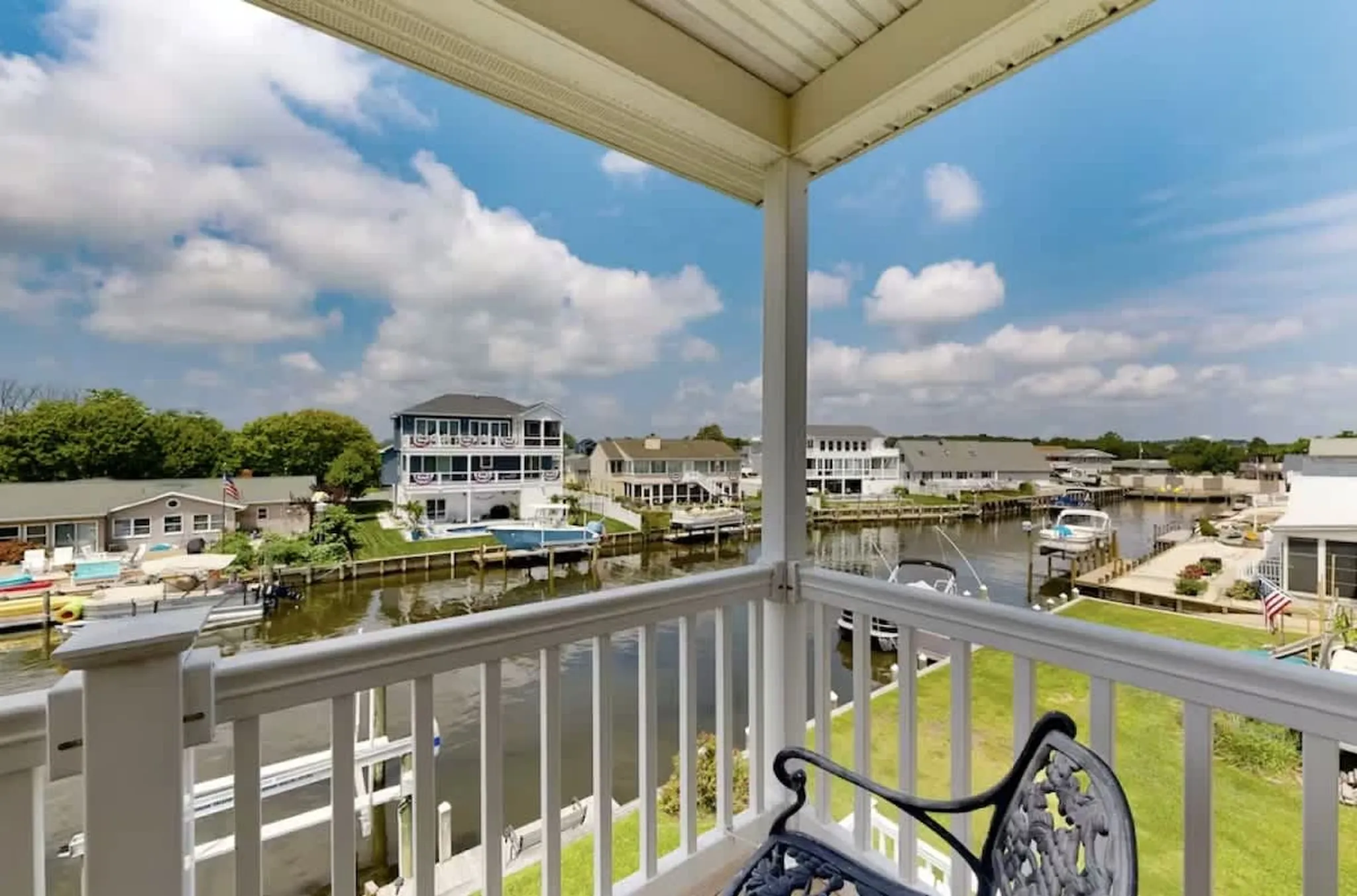Covered balcony with white railing and metal chair overlooking a canal lined with waterfront homes, docks and boats under blue sky