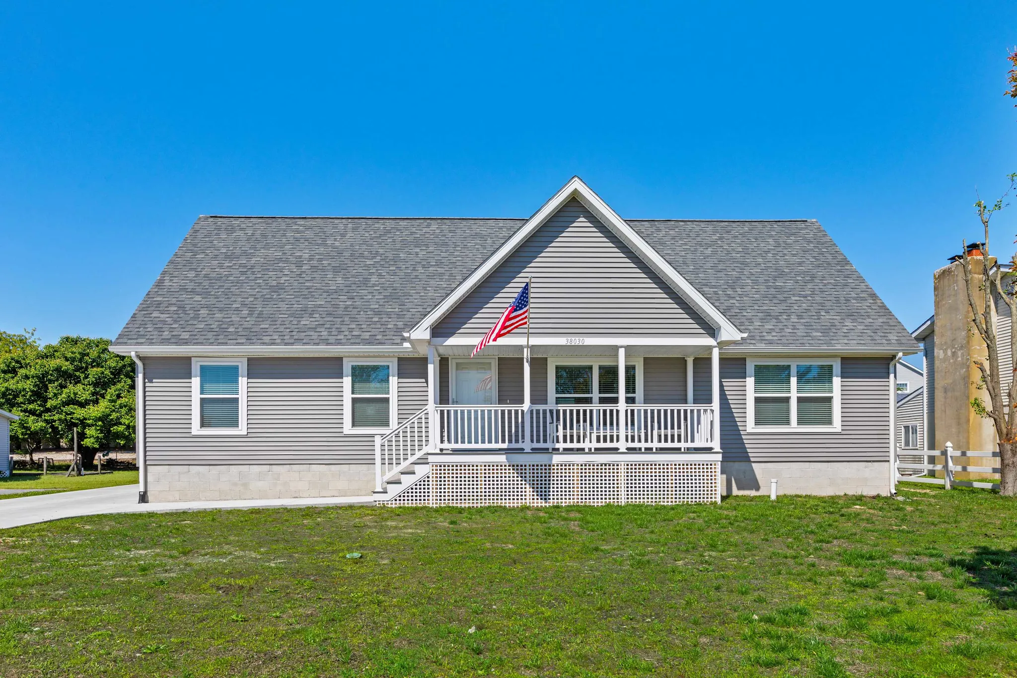 Single-story gray house with white front porch and American flag under clear blue sky, green lawn in foreground