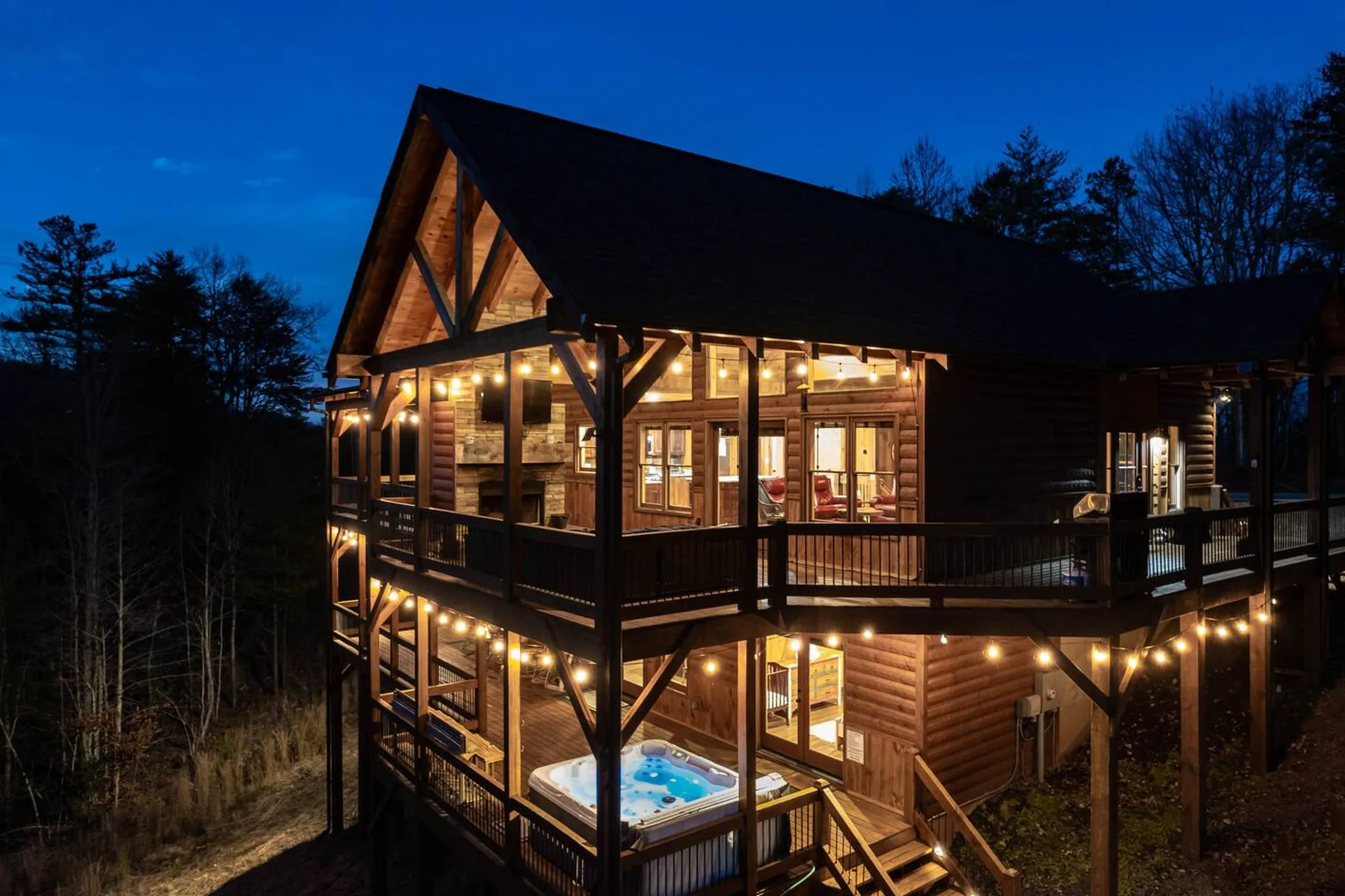 Two-story wood cabin at dusk with glowing string lights, wraparound decks, hot tub, and warm interior lights against a deep blue sky