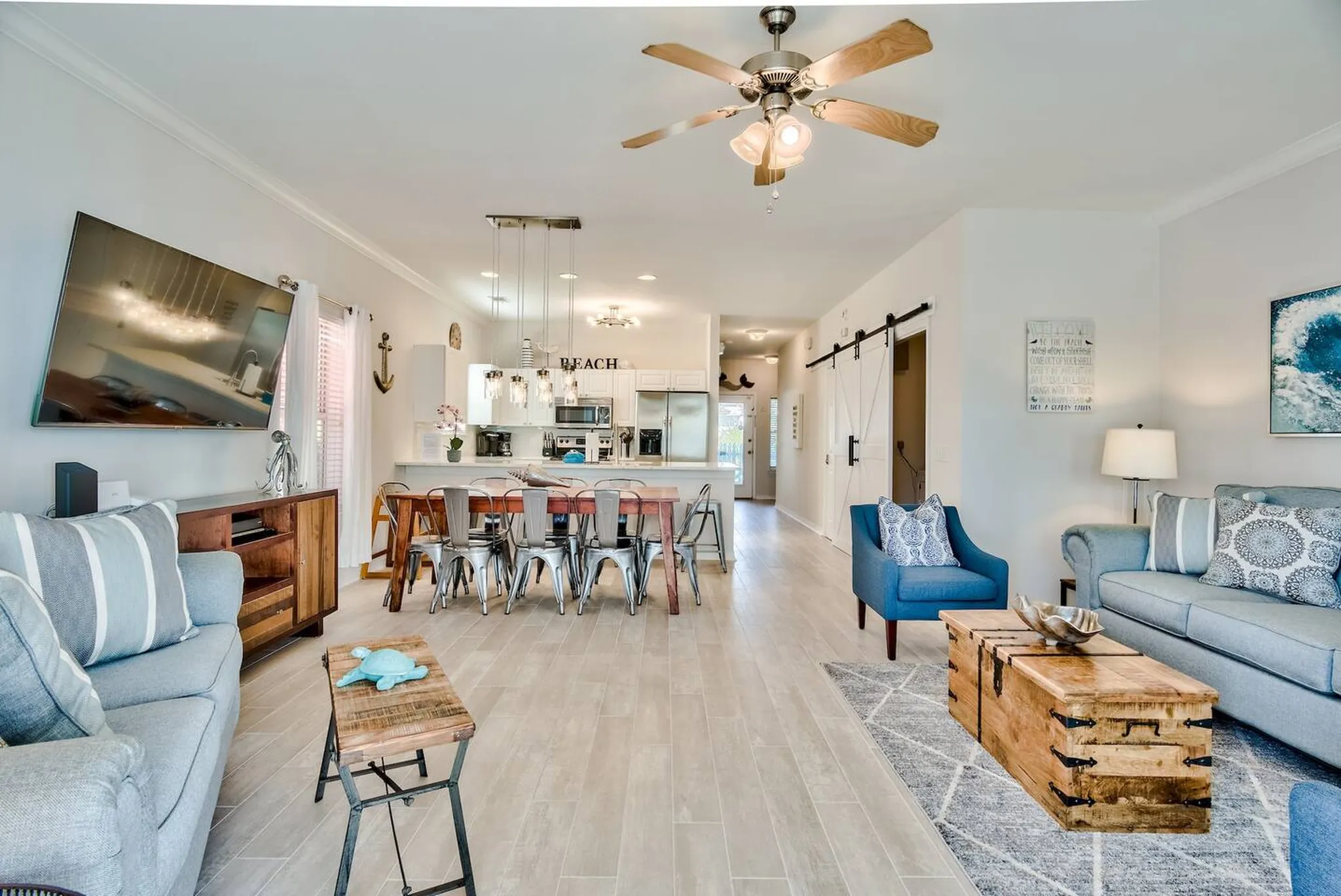 Open-concept coastal living and dining area with ceiling fan, wall-mounted TV, metal chairs around dining table, and barn door to kitchen