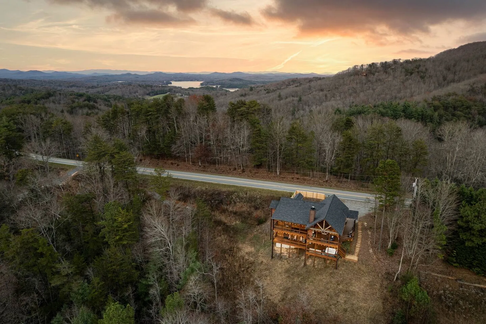 Aerial view of a wooden mountain cabin with wraparound decks surrounded by forest, with a lake and rolling hills at sunset