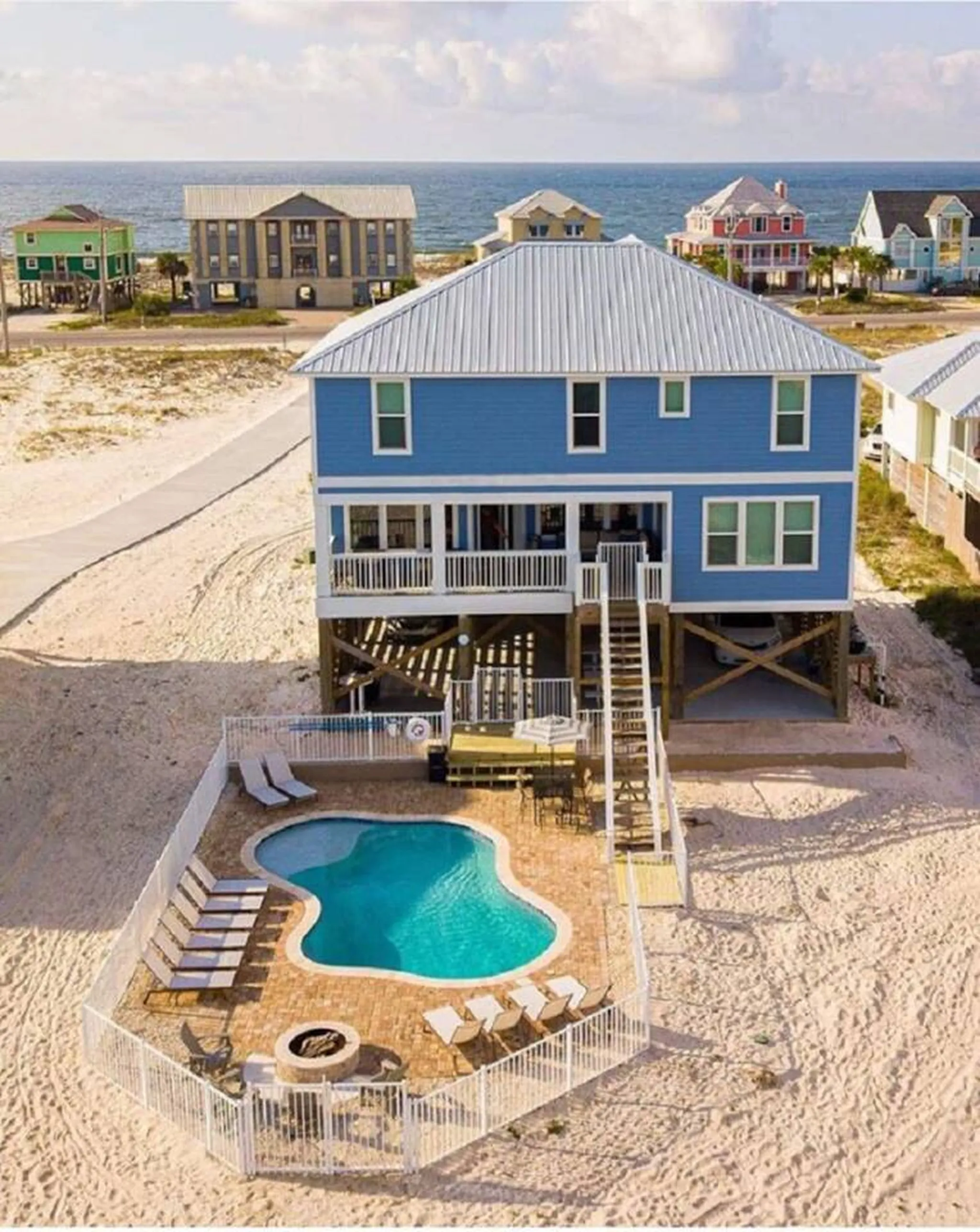 Aerial view of a blue elevated beach house with a kidney-shaped pool, lounge chairs, and fire pit on sandy coastline