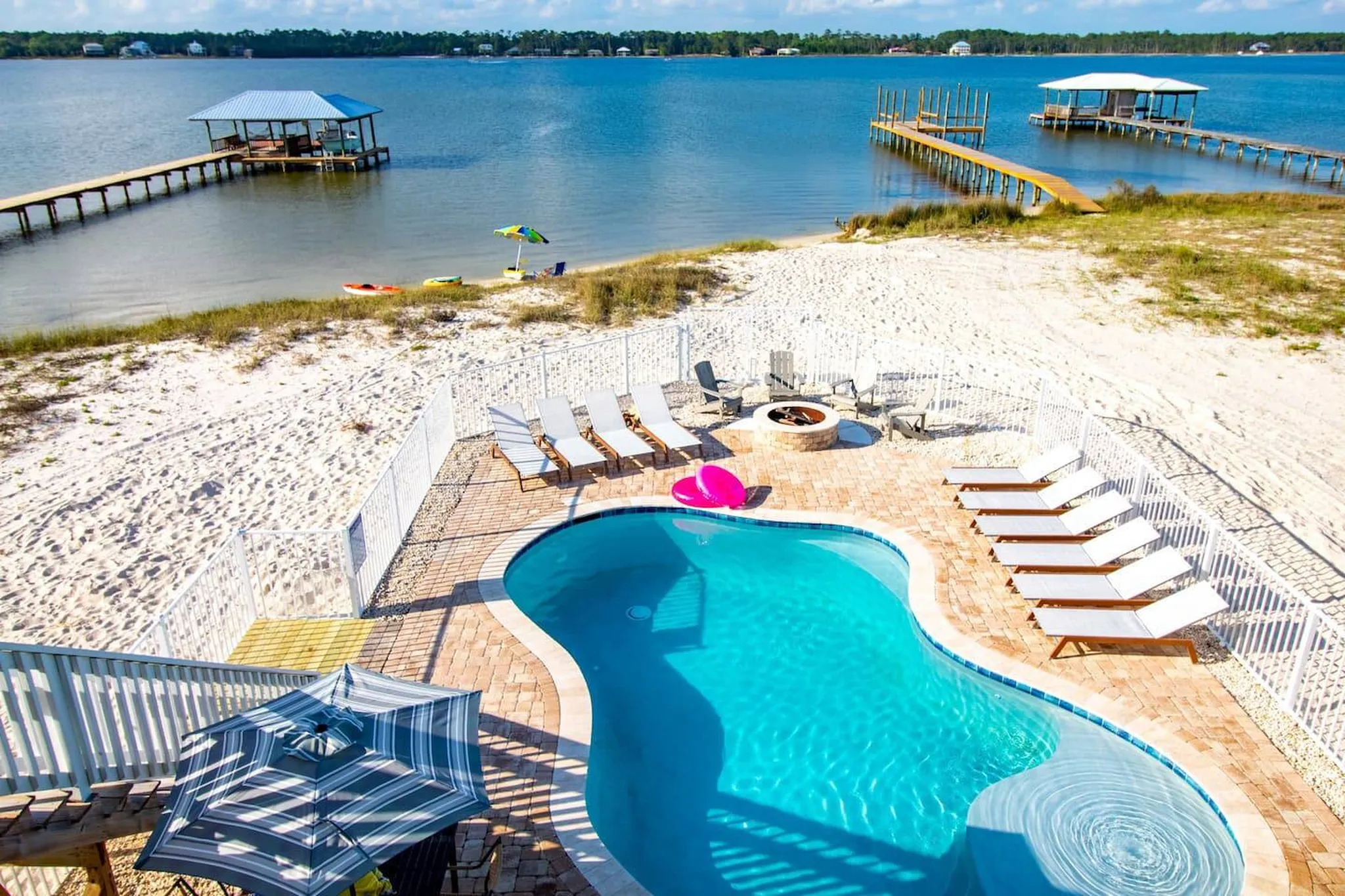 Waterfront backyard with curved pool, lounge chairs, fire pit, and private docks extending into a calm blue bay
