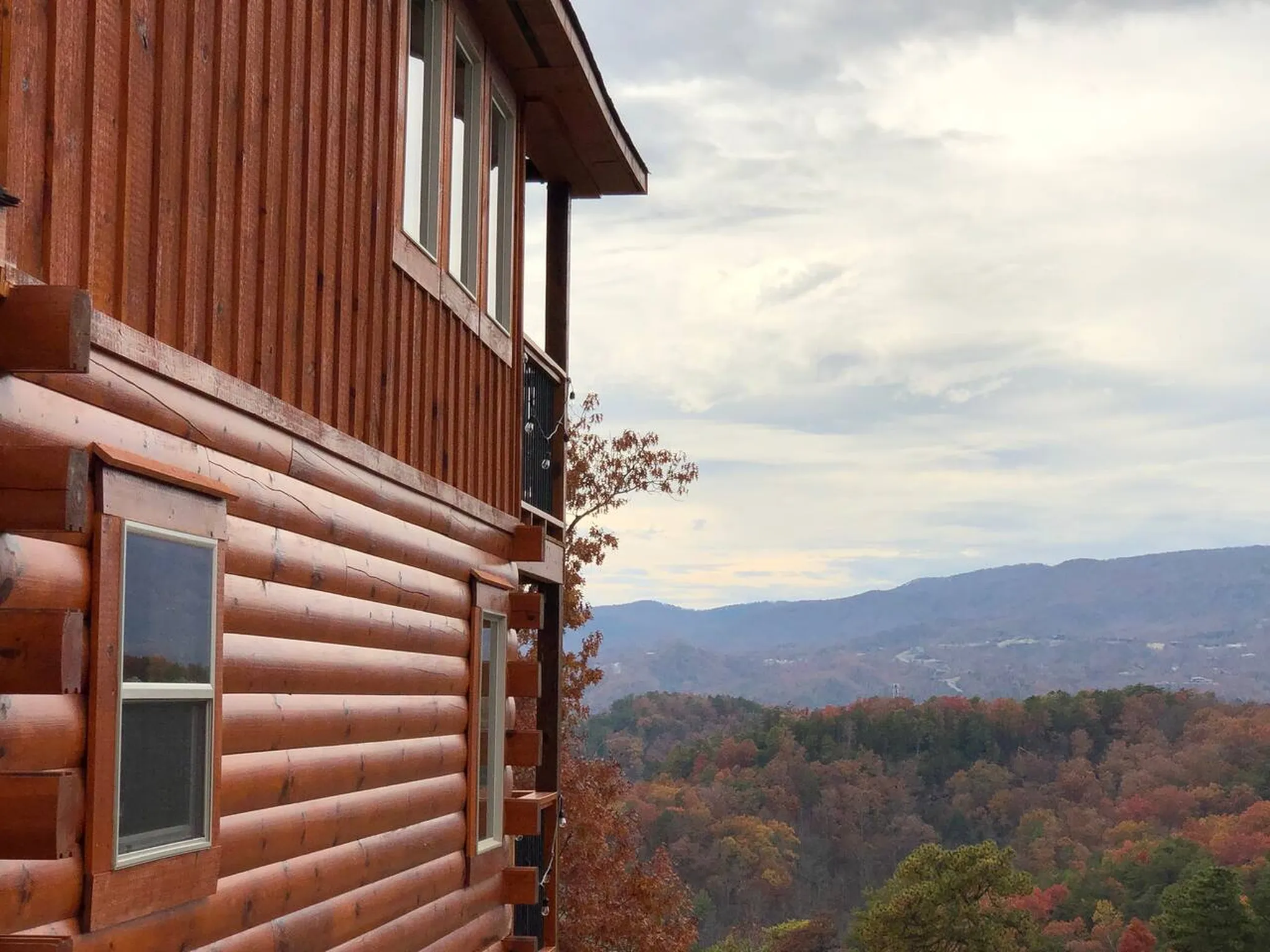 Log cabin exterior overlooking rolling forested mountains with autumn foliage under a partly cloudy sky