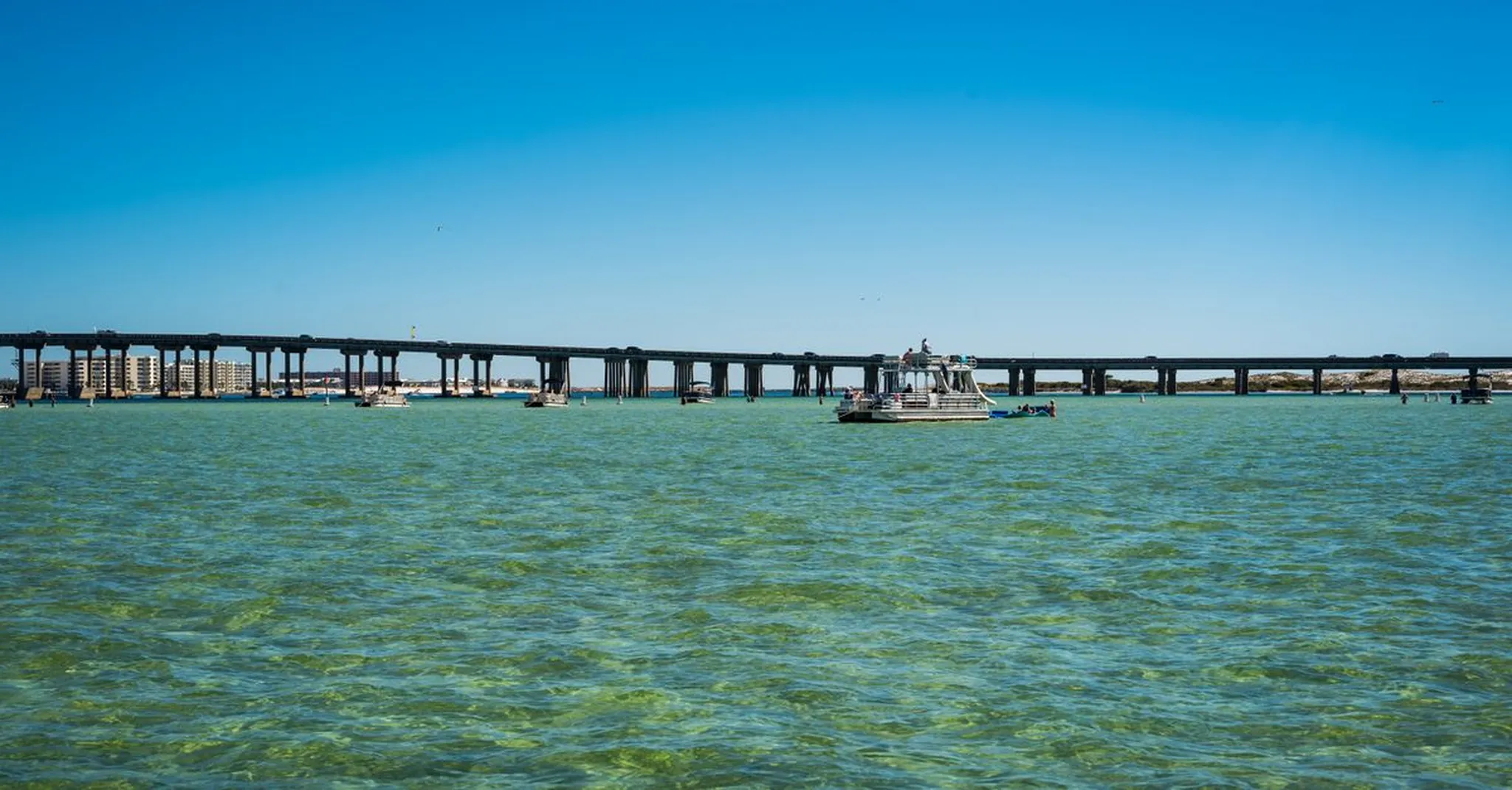 Long low bridge spanning turquoise coastal water with a small boat passing beneath and buildings visible on the far shore