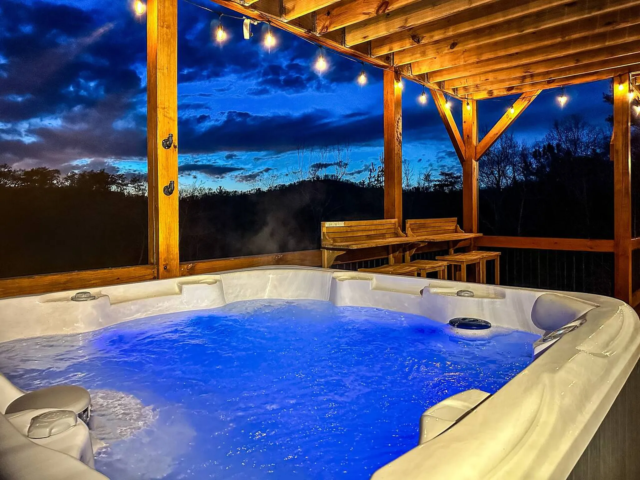 Steaming hot tub glowing blue on covered deck at dusk with string lights and mountain views