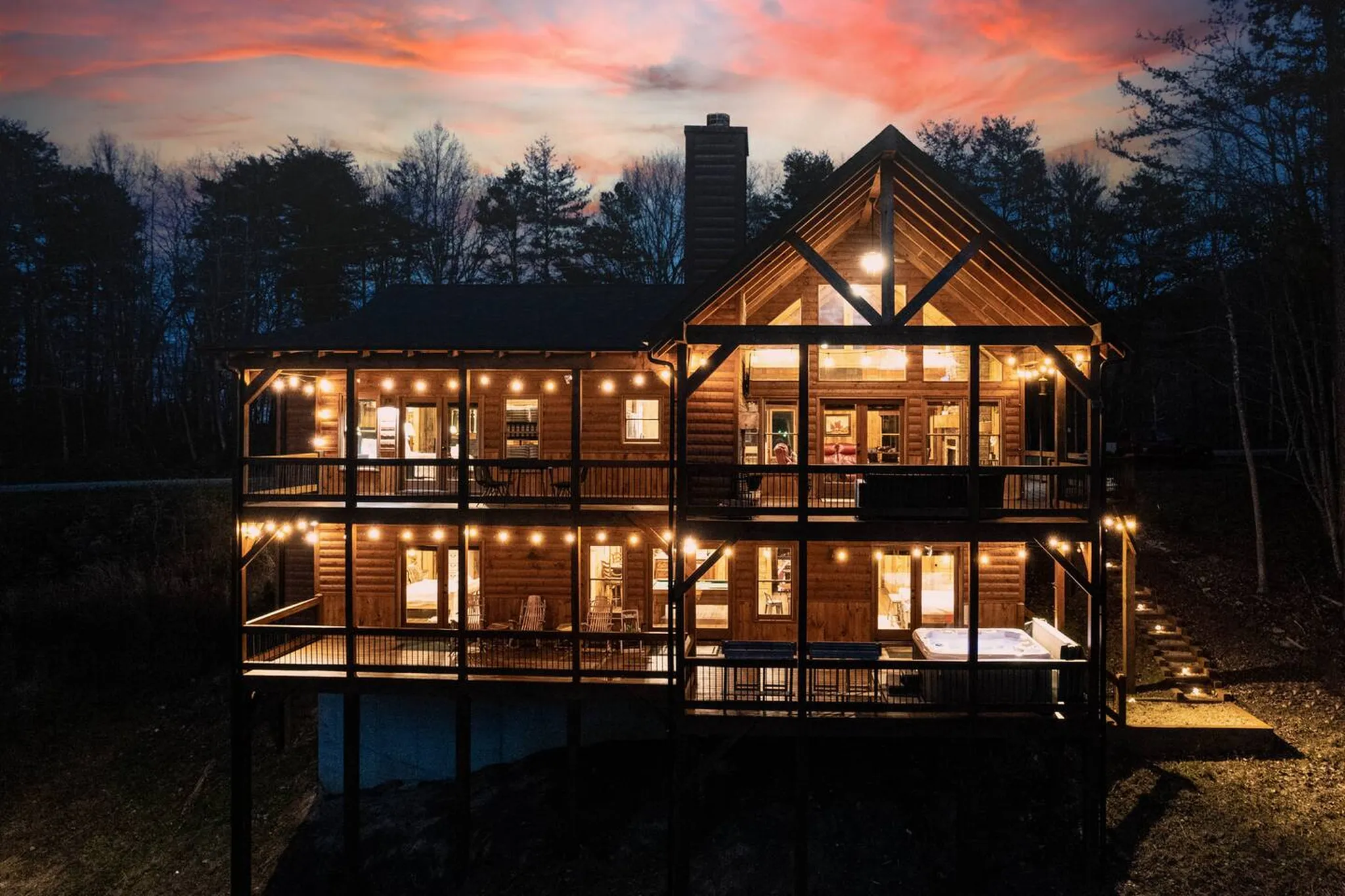 Multi-level wooden cabin at dusk with glowing string lights, wraparound decks, stone chimney, and a pink sunset sky over forested ridgeline