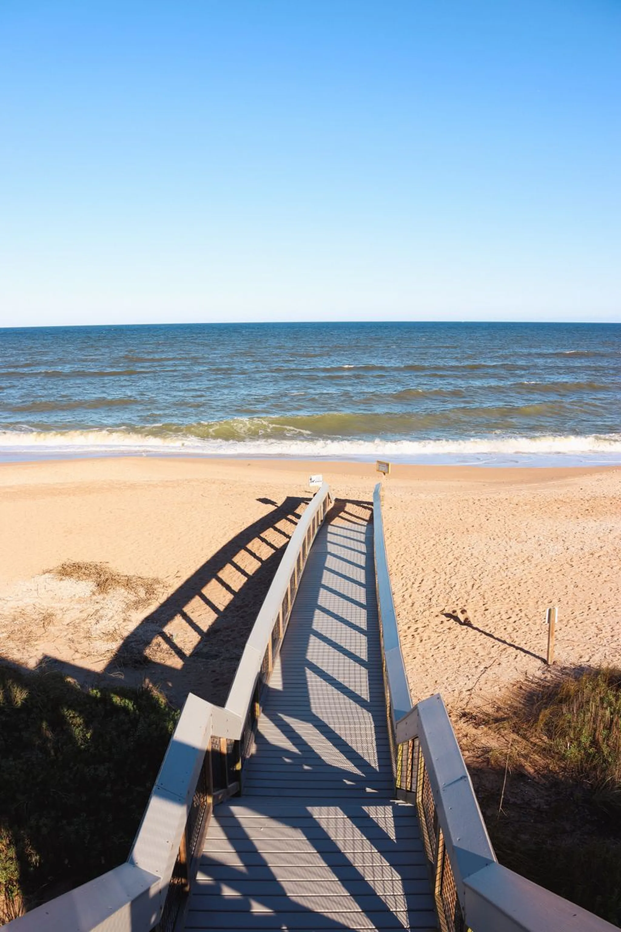 Wooden boardwalk stairs leading down to a sandy beach with gentle waves and clear blue sky