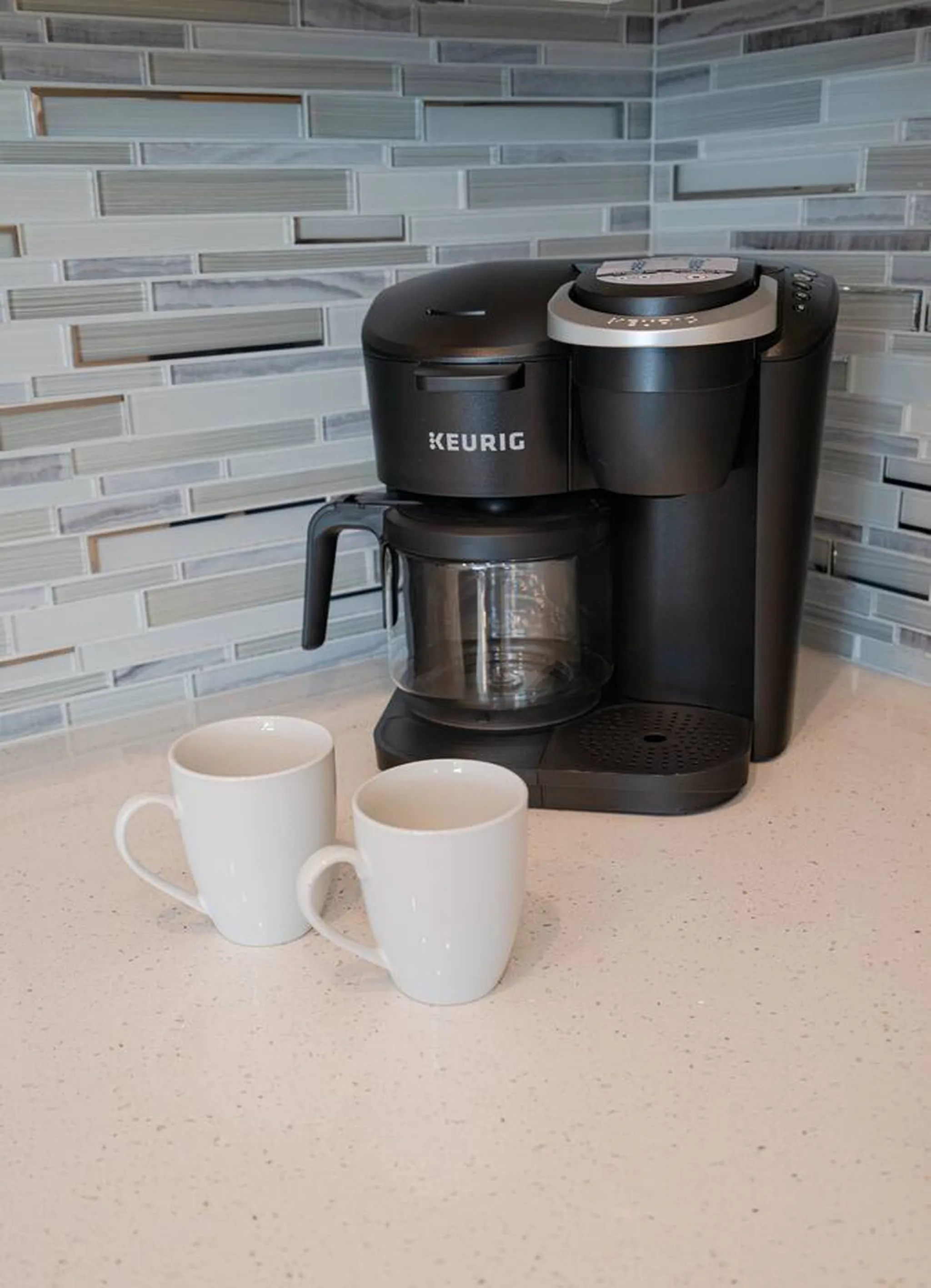 Keurig coffee maker with glass carafe and two white mugs on a quartz countertop against a blue glass tile backsplash