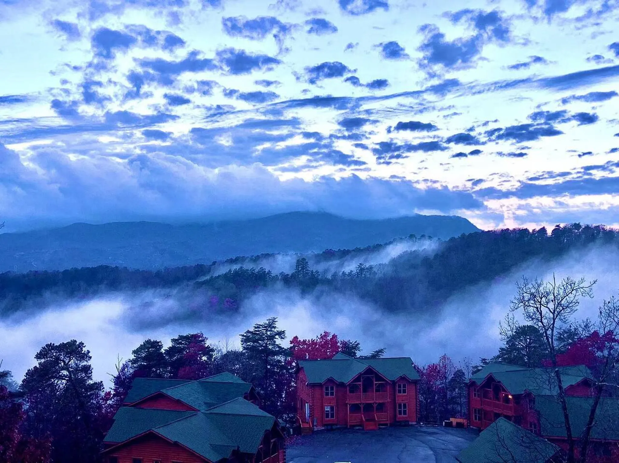 Wooden cabins on a forested ridge at dusk with mist rolling through blue mountain valleys under a cloud-streaked sky