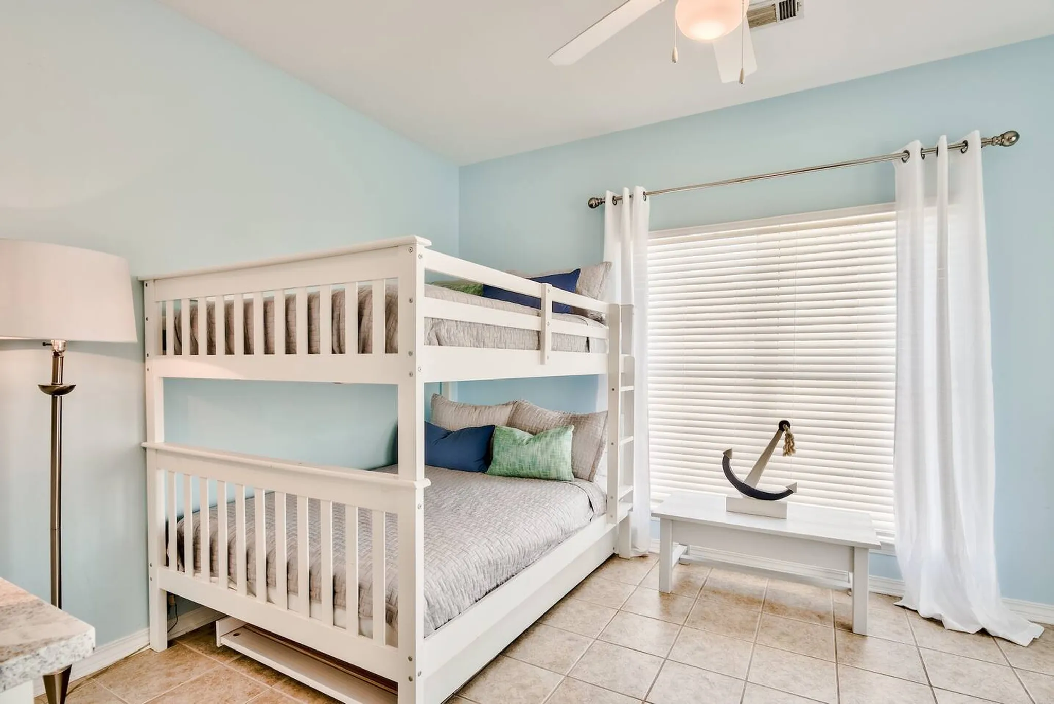 Light blue bedroom with white full-over-full bunk beds, ceiling fan, tile floor, and a wooden bench holding an anchor decoration