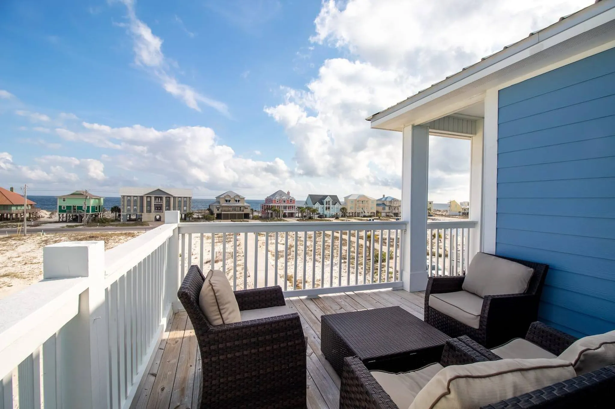 Beachfront balcony with wicker seating overlooking sandy shore and colorful coastal homes under a bright blue sky