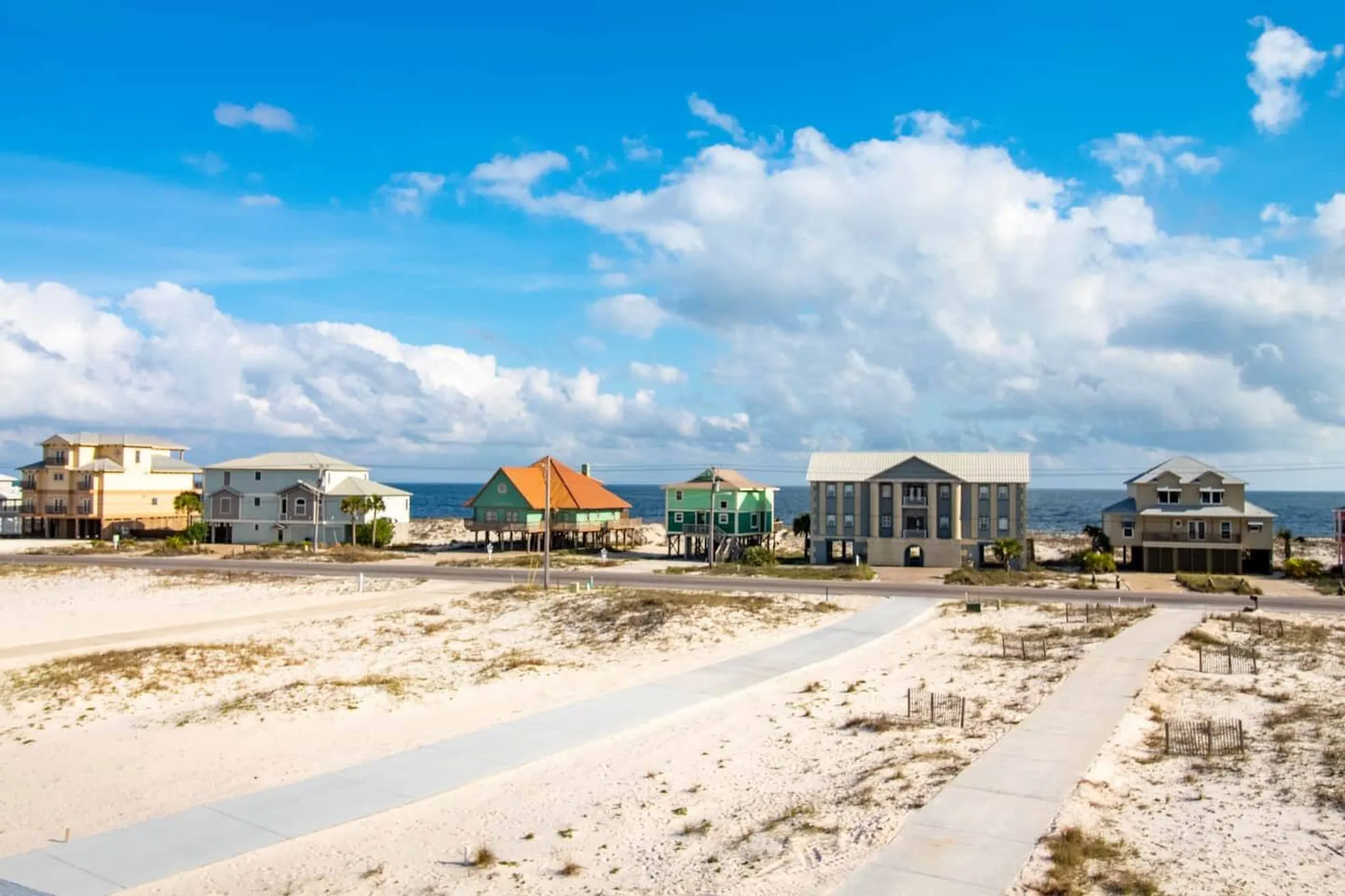 Row of colorful stilted beach houses along a sandy coastline under a bright blue sky with scattered clouds