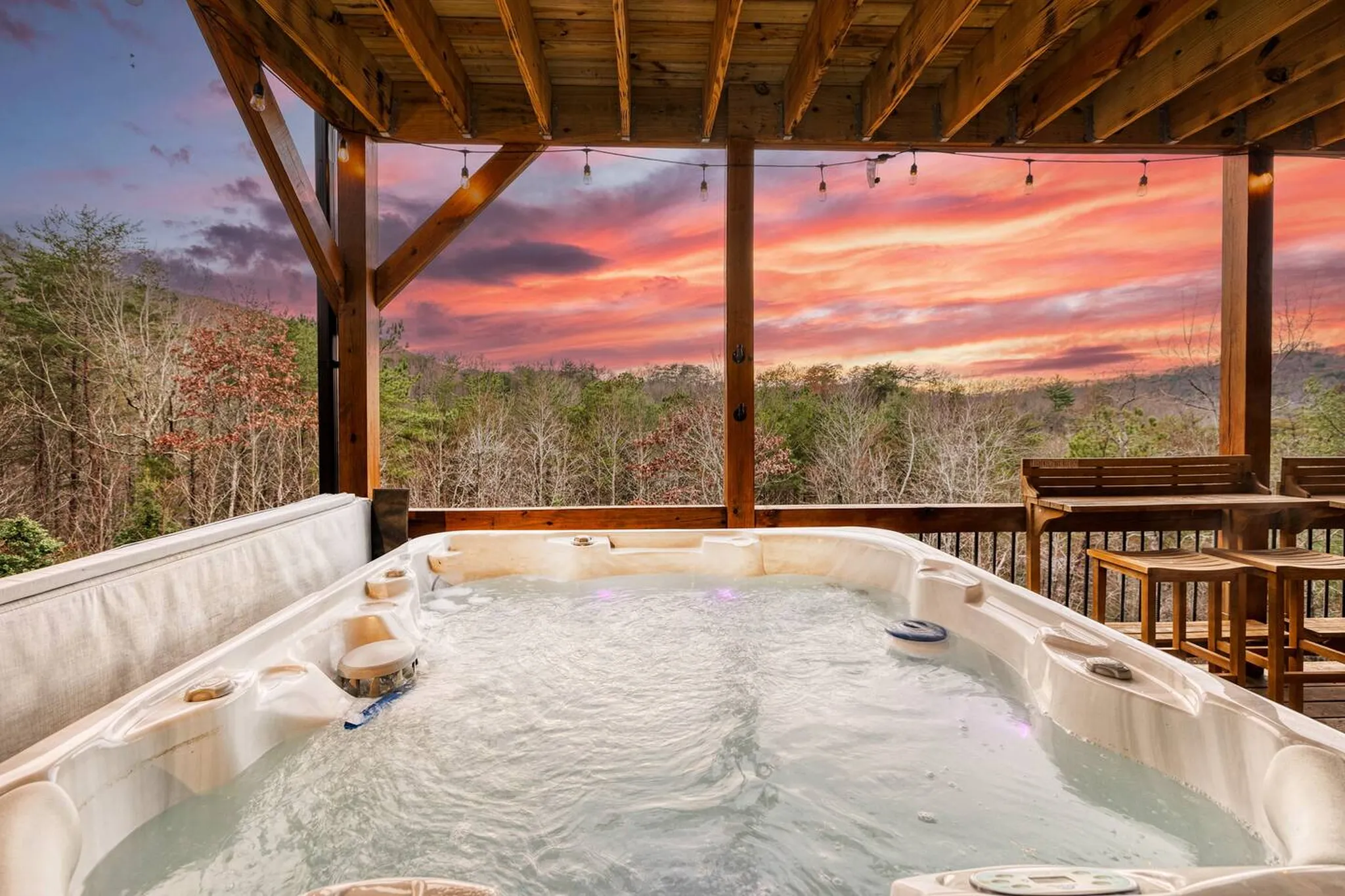 Bubbling hot tub on covered wooden deck with string lights overlooking forested mountains at sunset with pink and orange sky
