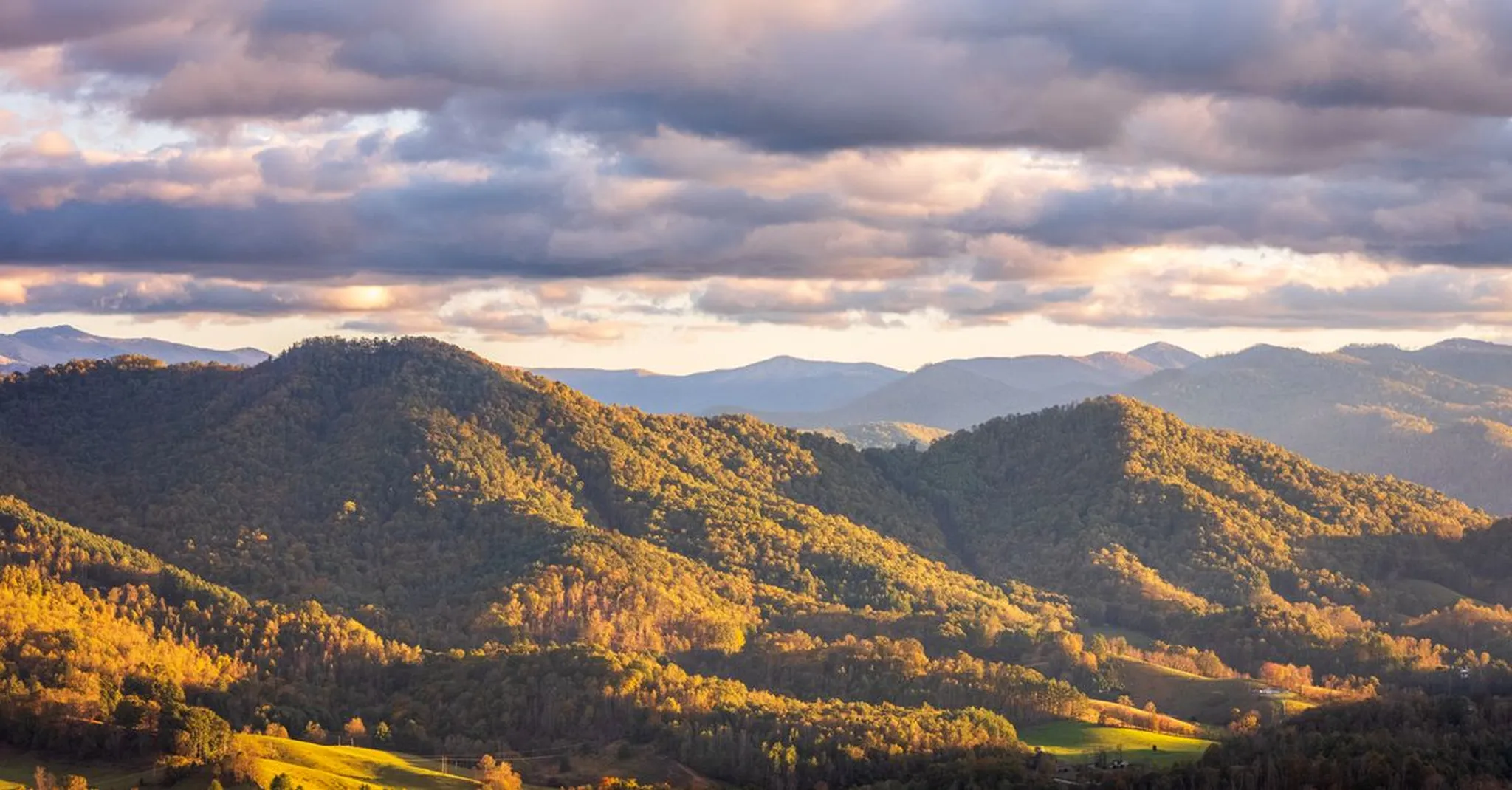 Rolling forested mountains bathed in golden sunset light under a dramatic cloudy sky, with layered ridges fading into the distance