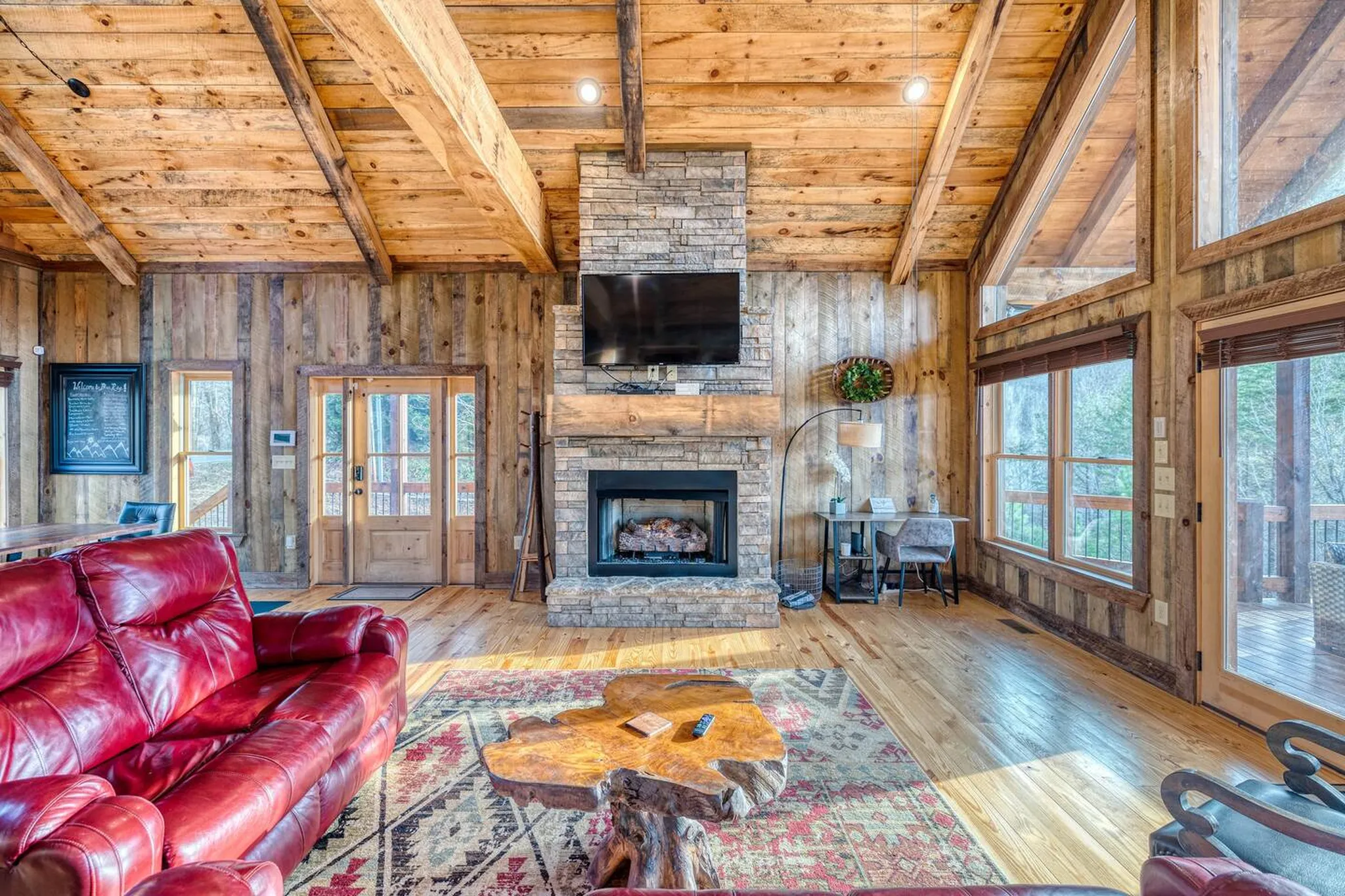 Rustic wood-beamed living room with red leather sofa, stone fireplace, mounted TV, and live-edge coffee table on patterned rug