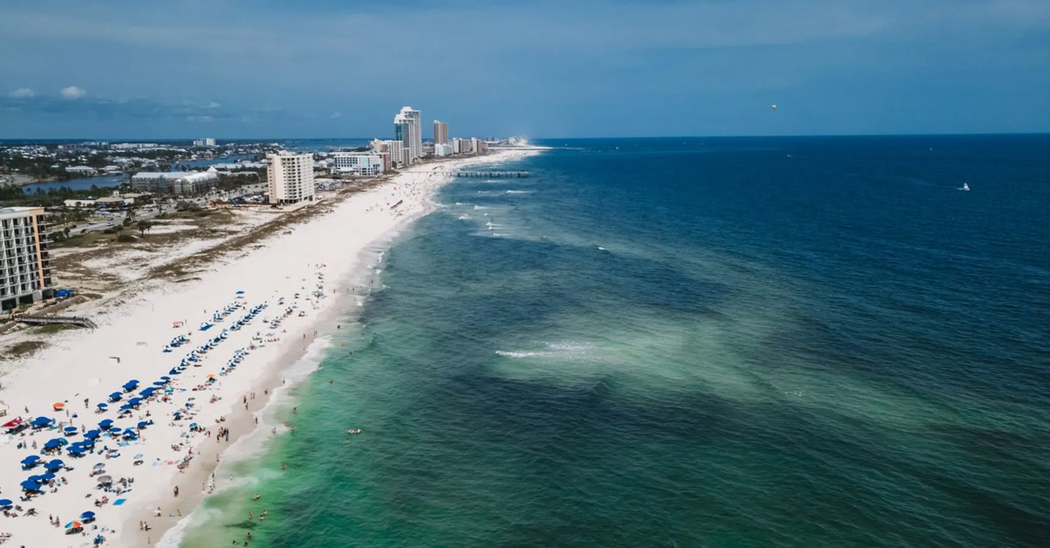 Aerial view of a long sandy beach lined with colorful umbrellas, high-rise condos, and turquoise ocean water under a cloudy sky