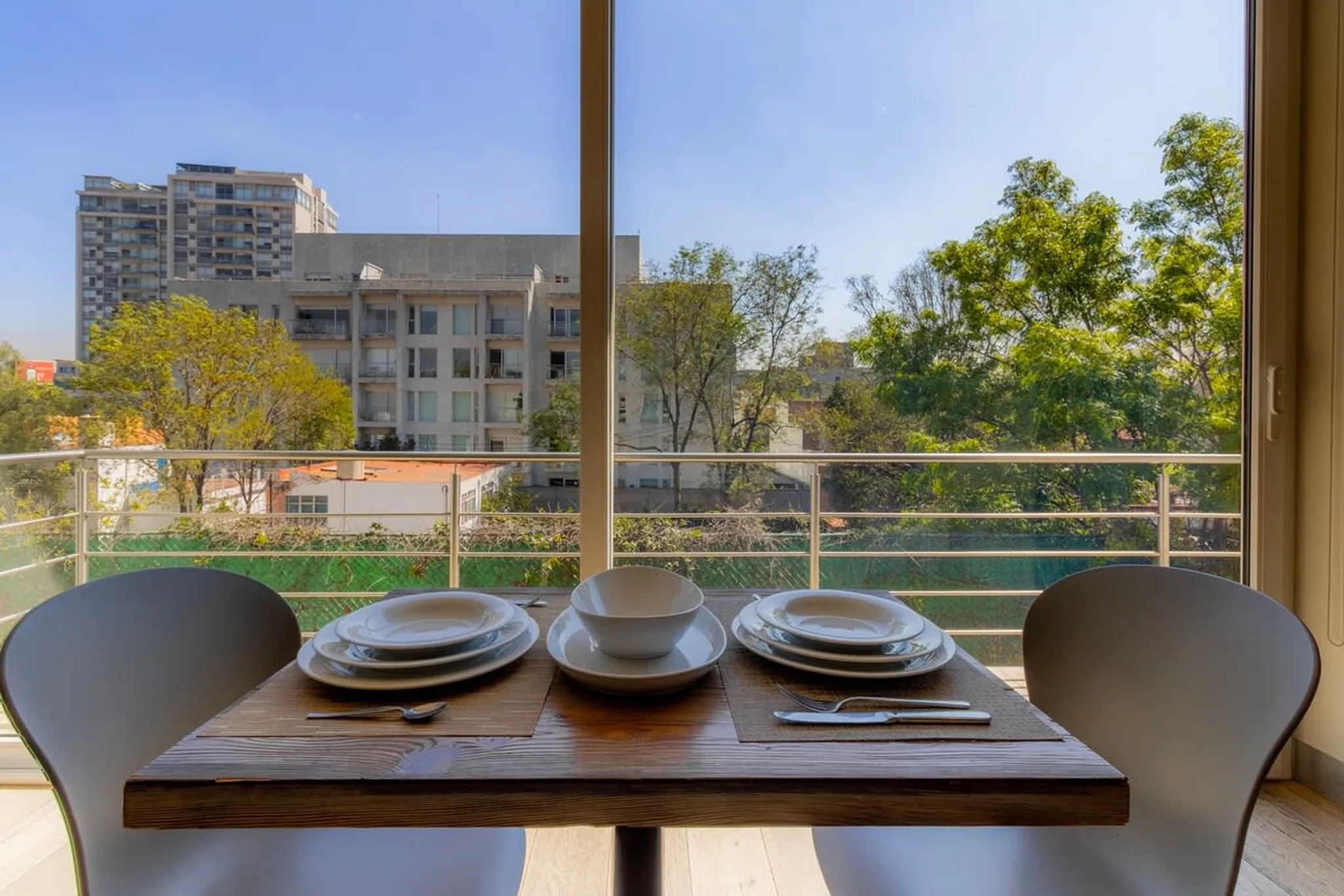 A dining table with place settings is set on a balcony with a view of apartment buildings and trees.