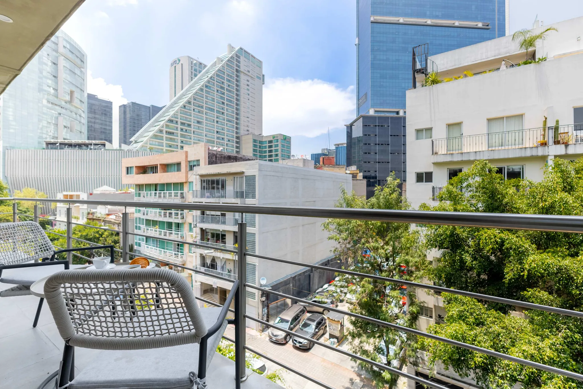 A balcony with chairs and a table overlooking a city with buildings and trees.