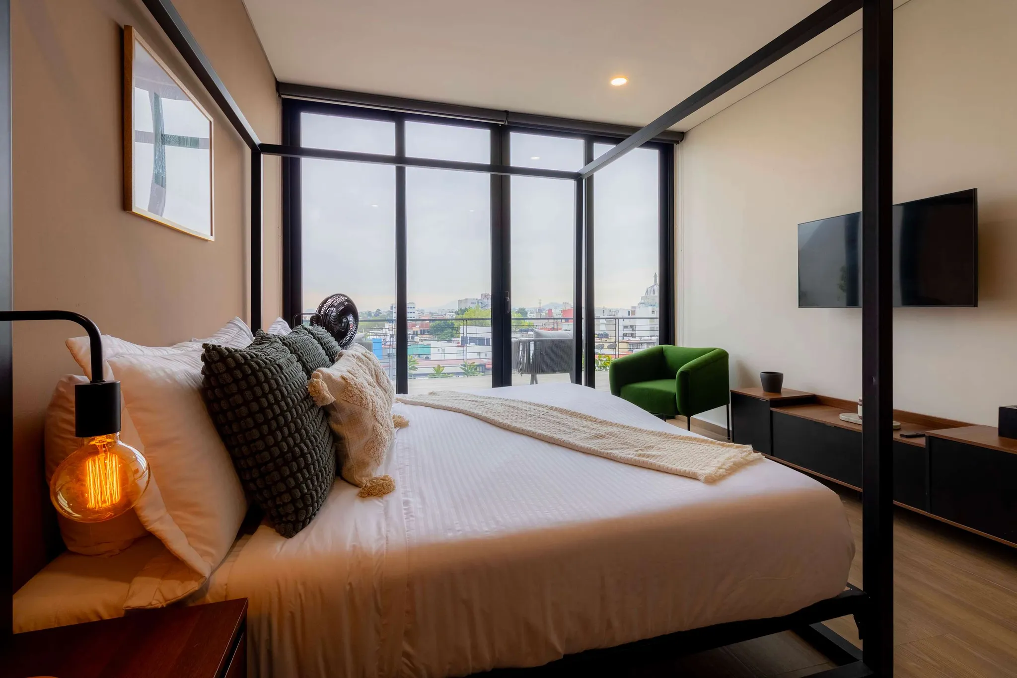 A bedroom with a canopy bed, green armchair, and large windows overlooking a city.