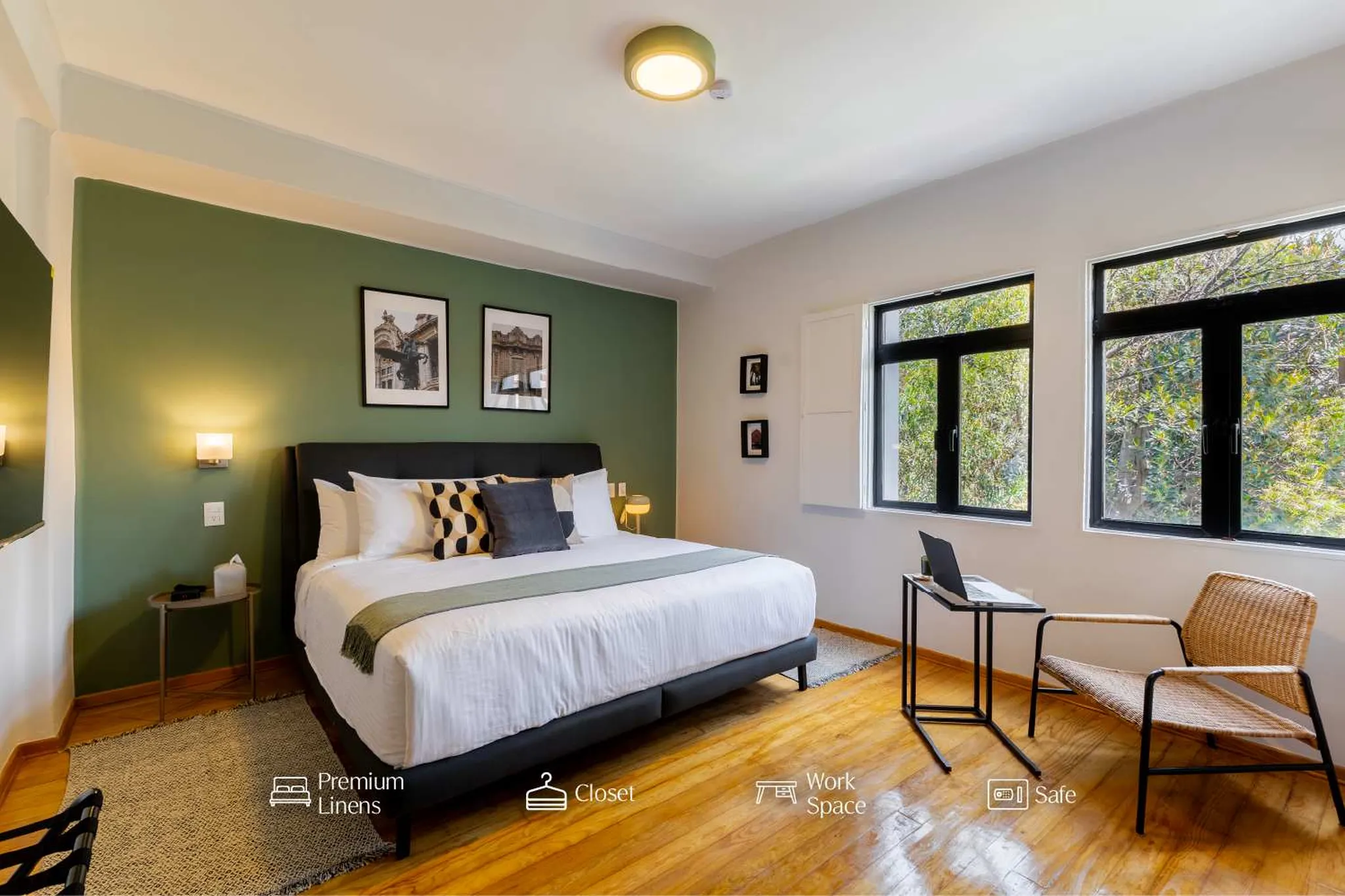 A stylish bedroom with a green accent wall, large bed, and two windows overlooking lush trees.