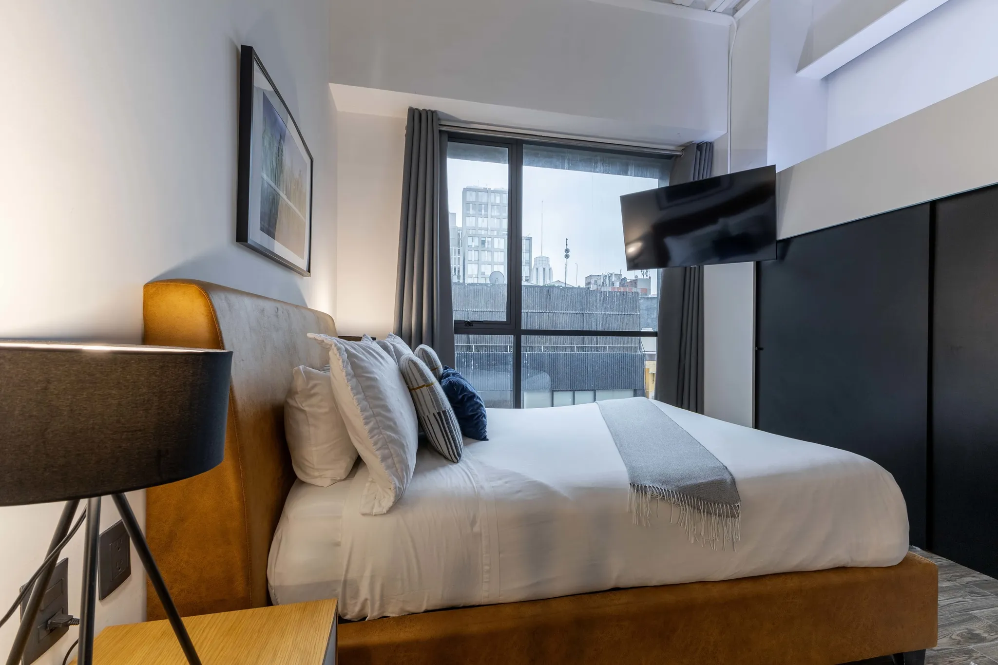A king-sized bed with a mustard-colored headboard and white linens sits near a window with a city vi