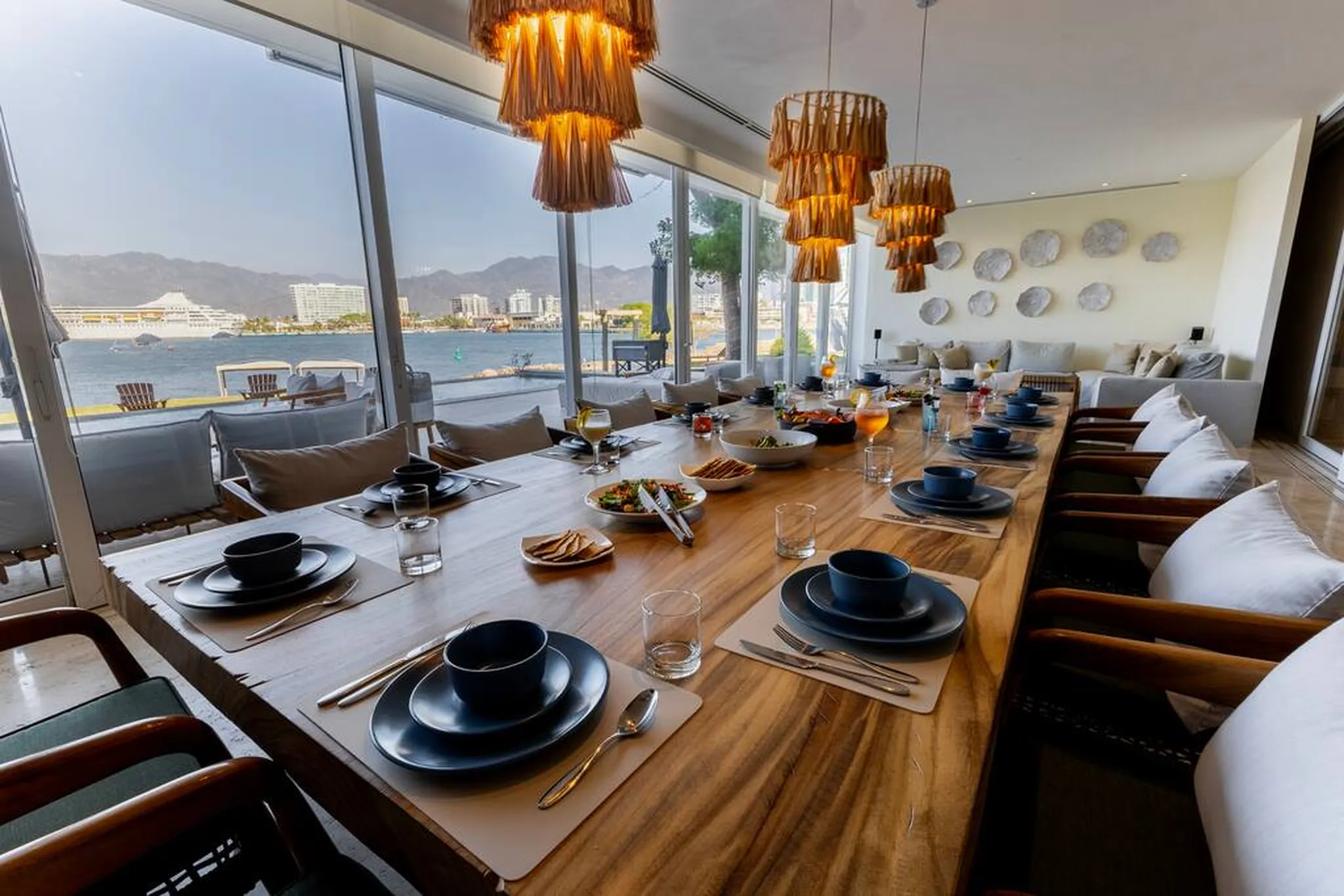 A long wooden dining table is set with dark blue plates and bowls overlooking the ocean.