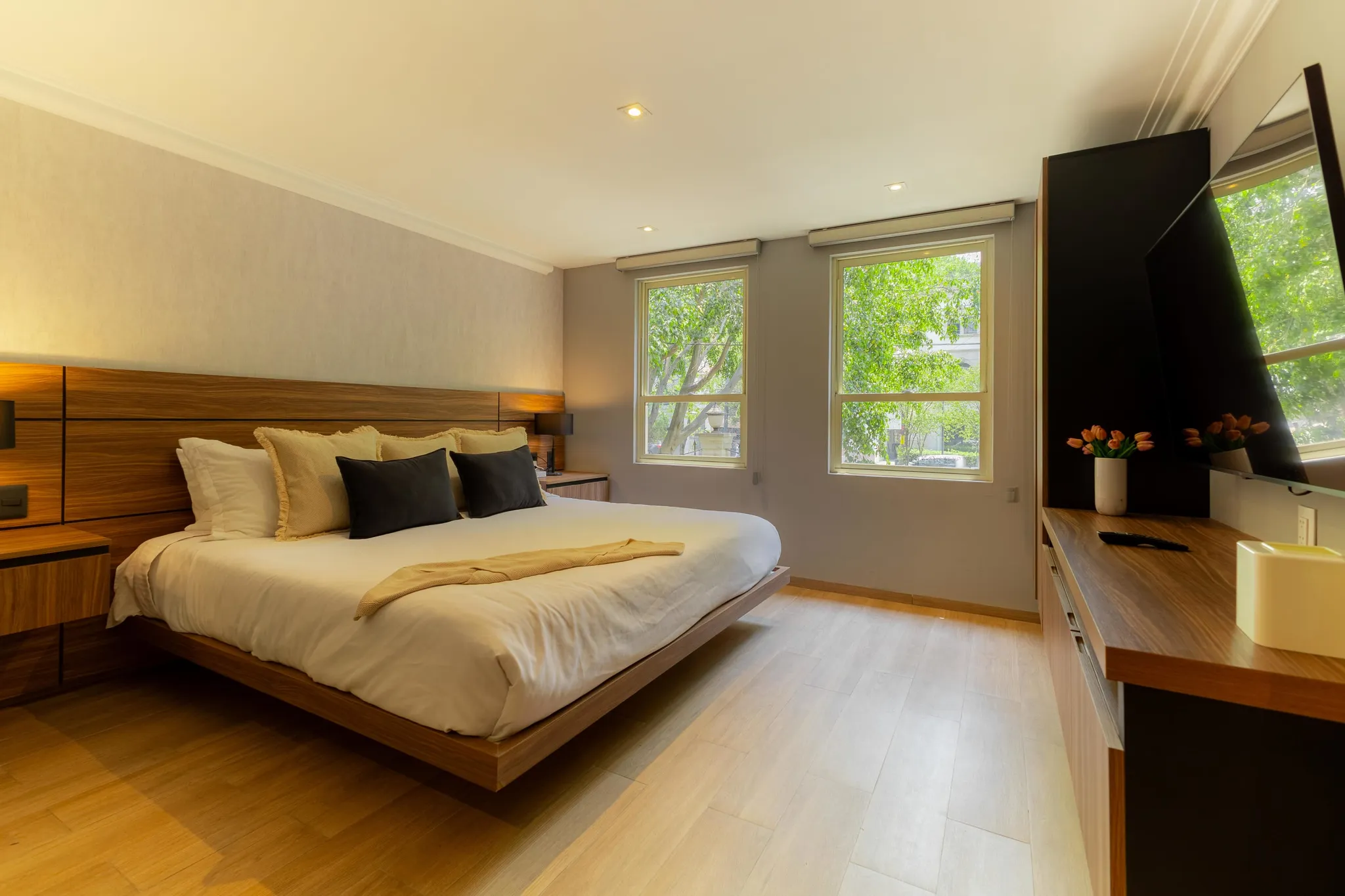Modern bedroom with a king-size bed, wooden headboard, and windows overlooking trees.