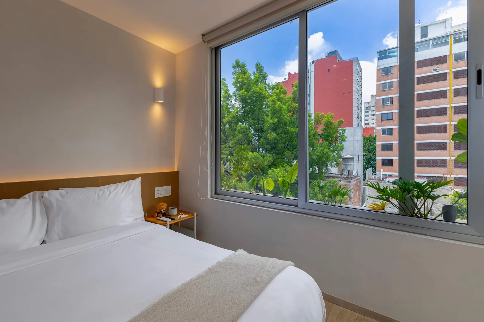 A neatly made bed with white linens sits next to a nightstand and a large window overlooking city bu