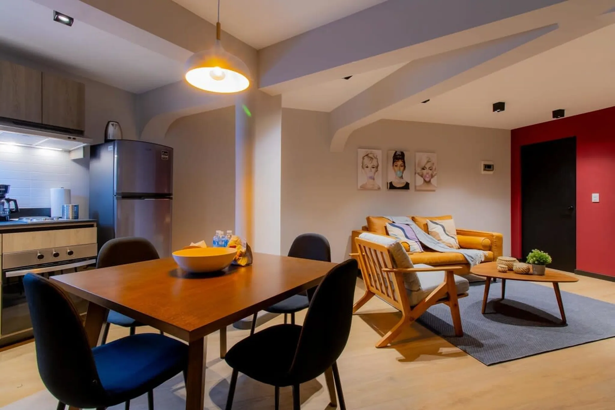 A dining table with chairs is in front of a kitchen with a refrigerator and stove.
