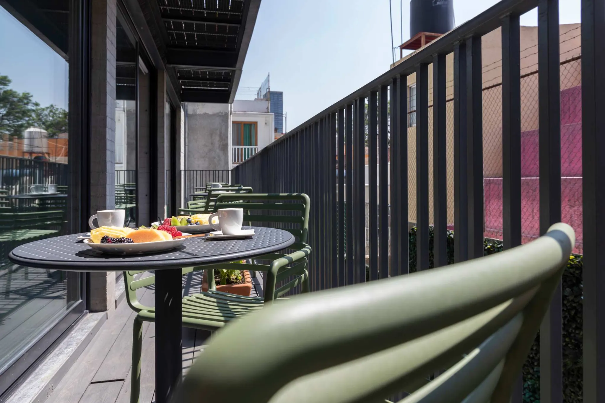 A table with fruit and coffee cups on a balcony with green chairs and a metal railing.