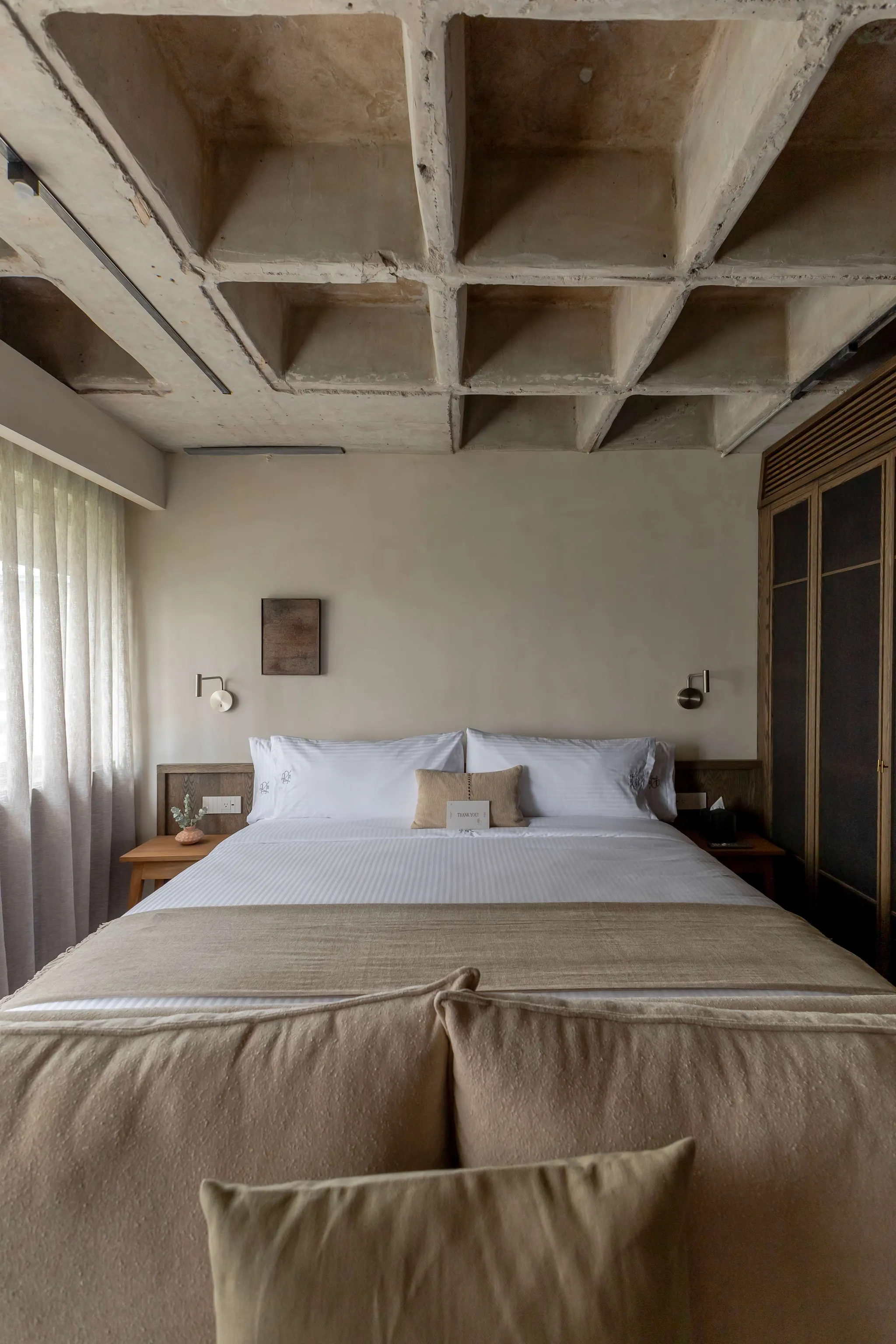 A minimalist hotel room with a concrete ceiling, bed, and curtains.
