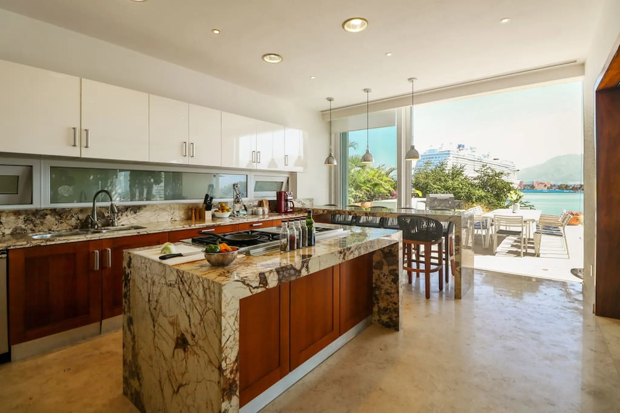 A modern kitchen with marble countertops and a large island opens to a patio with a view of a cruise