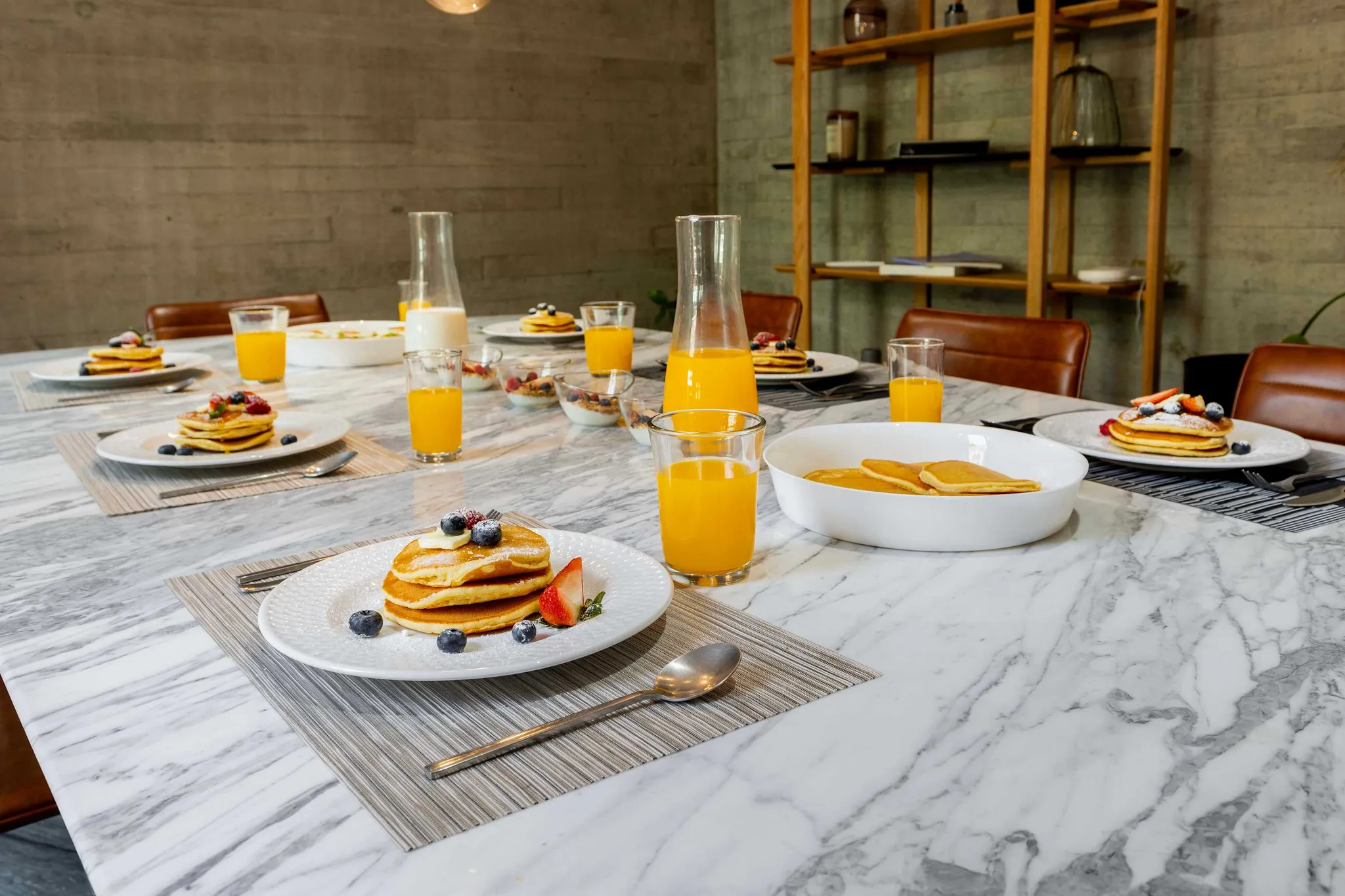 A marble table is set for breakfast with pancakes, fruit, juice, and milk.