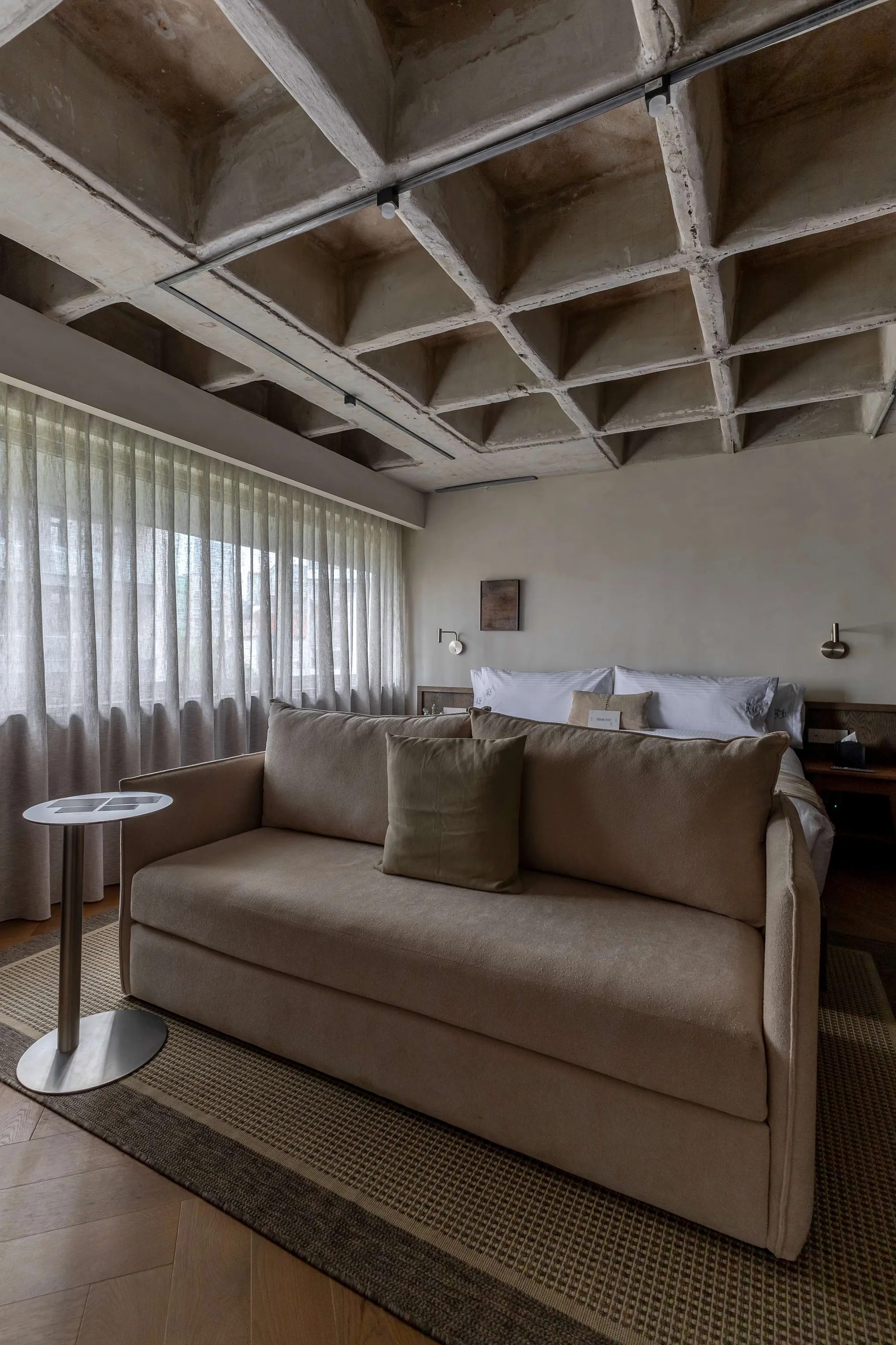 Modern hotel bedroom with a concrete coffered ceiling, sheer curtains, a couch, and a bed.