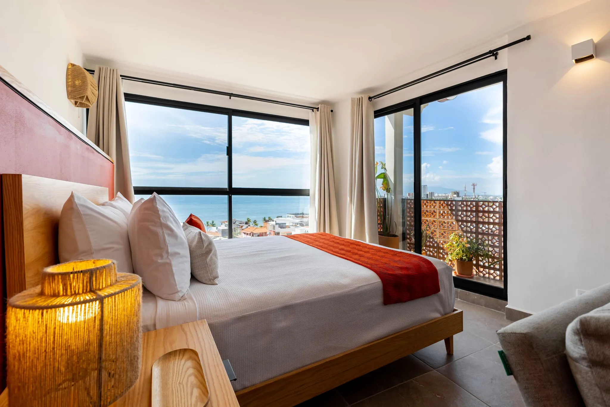 A king-size bed with white linens and an orange runner faces a window overlooking the ocean and city