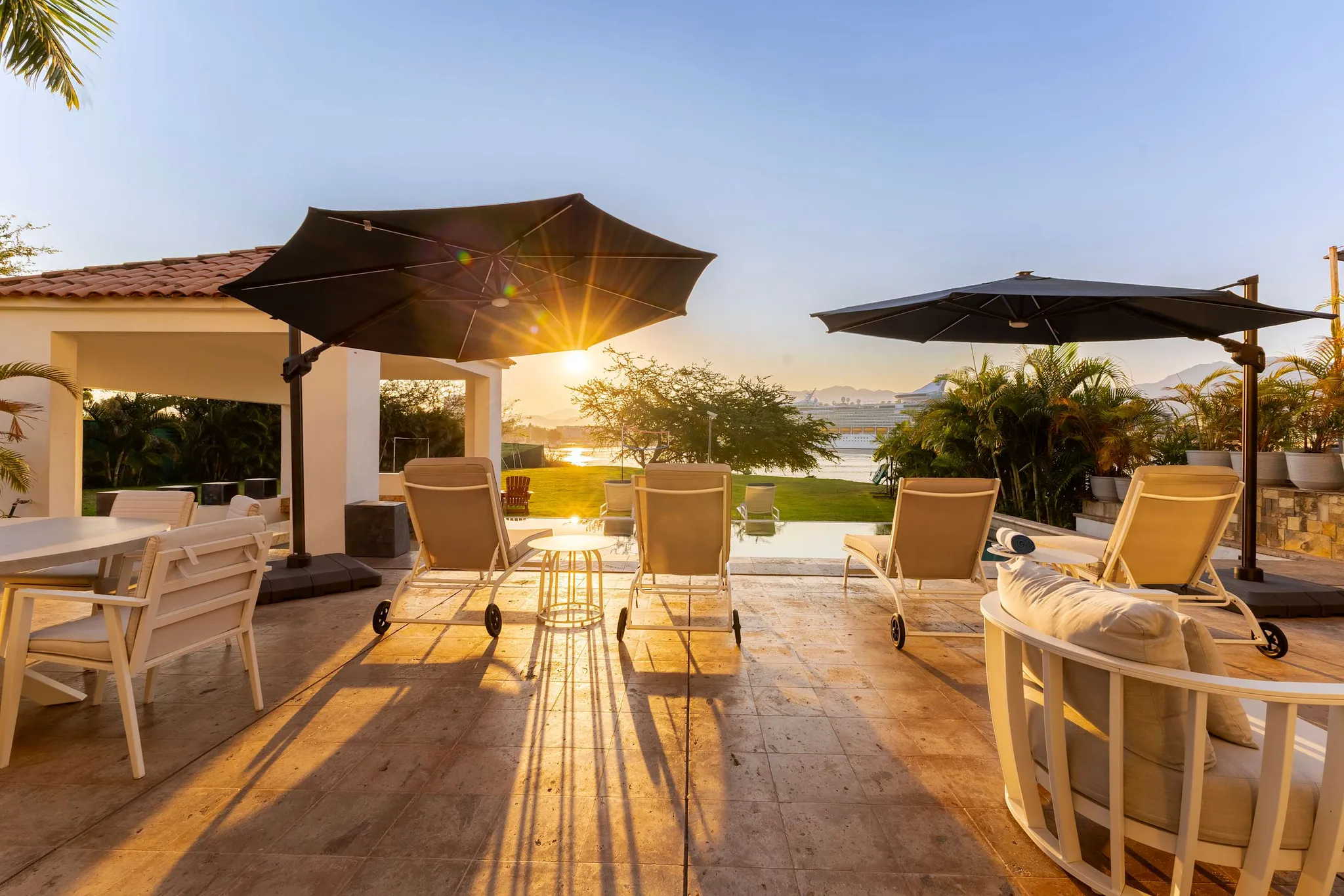Sunset casts long shadows on a patio with lounge chairs and umbrellas, overlooking a body of water w