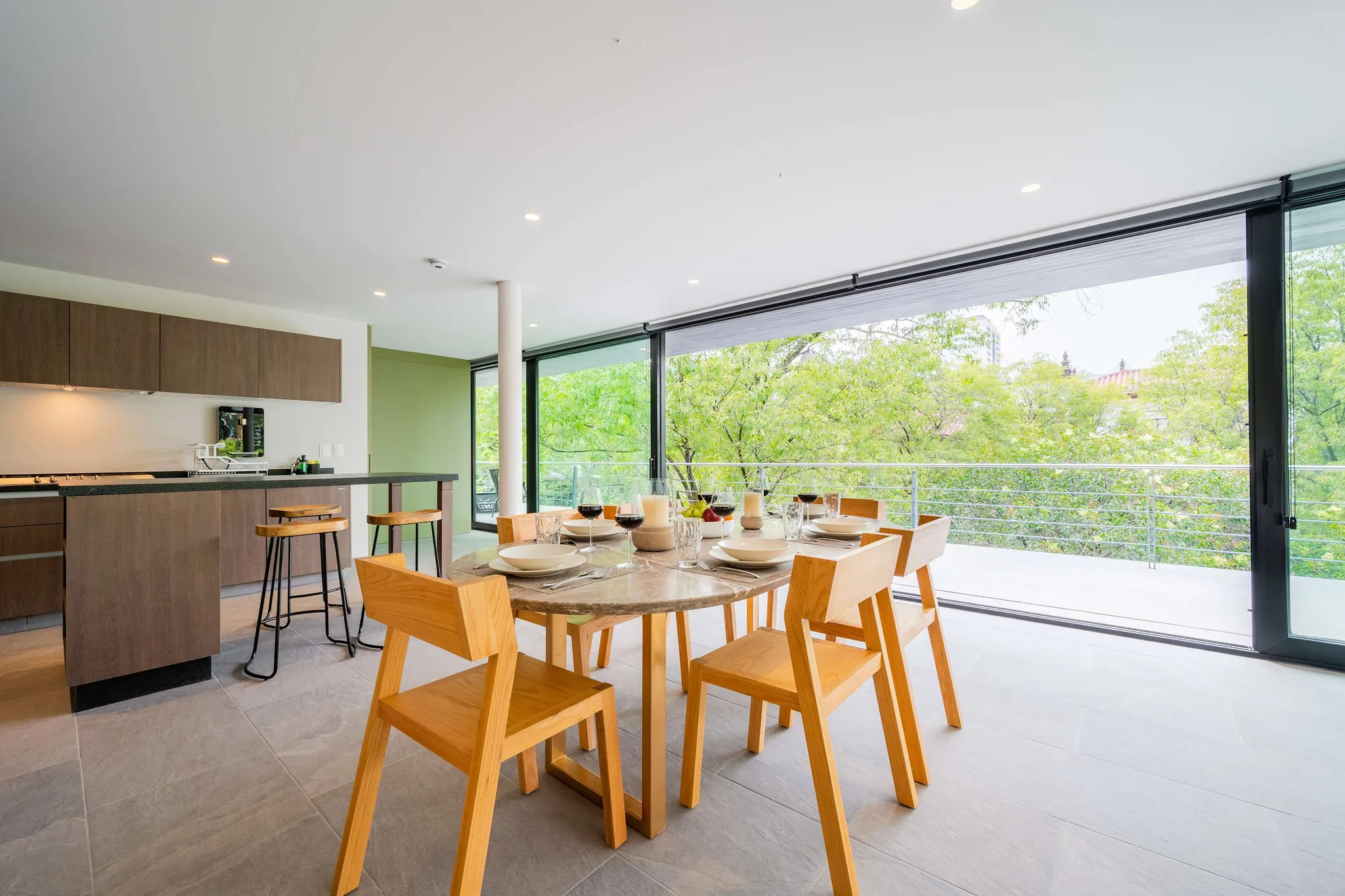 A modern dining room with a marble table, wooden chairs, and large glass doors with a view of trees.