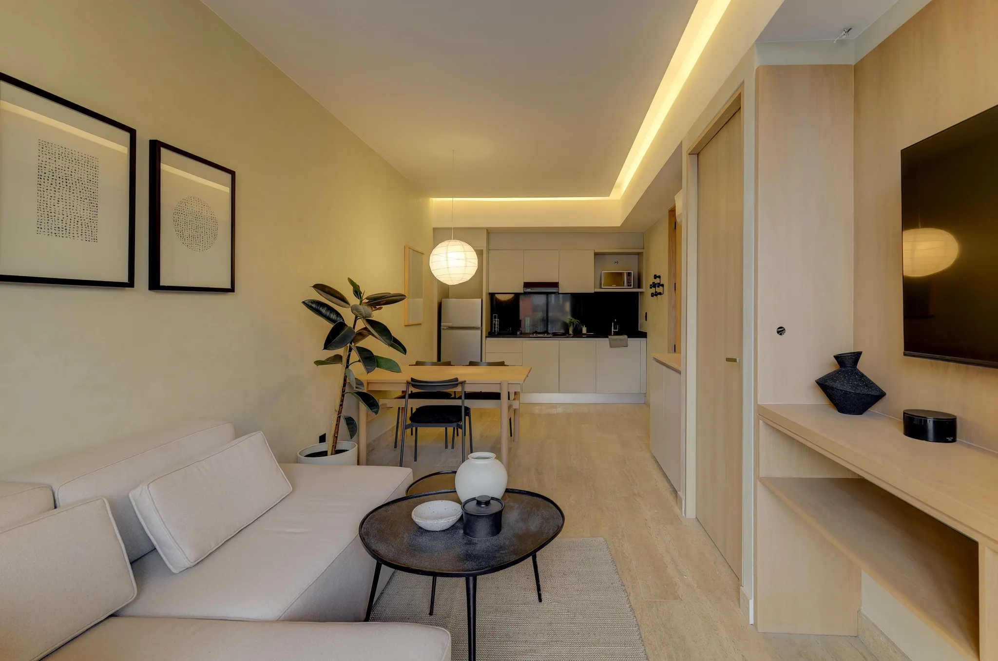 A bright, modern living room with a sectional sofa, coffee table, dining table, kitchen, and TV.