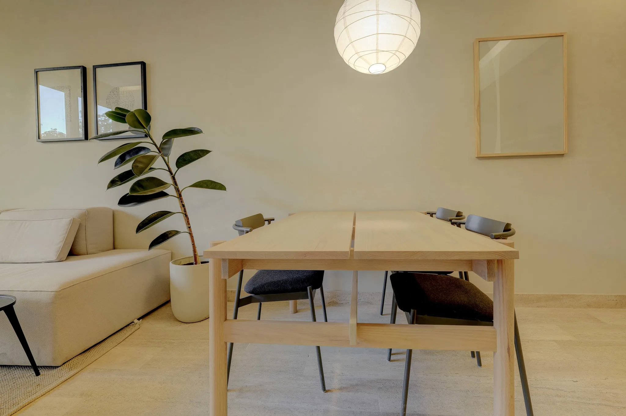 A modern dining room with a light wood table, minimalist chairs, a sectional sofa, and abstract art.