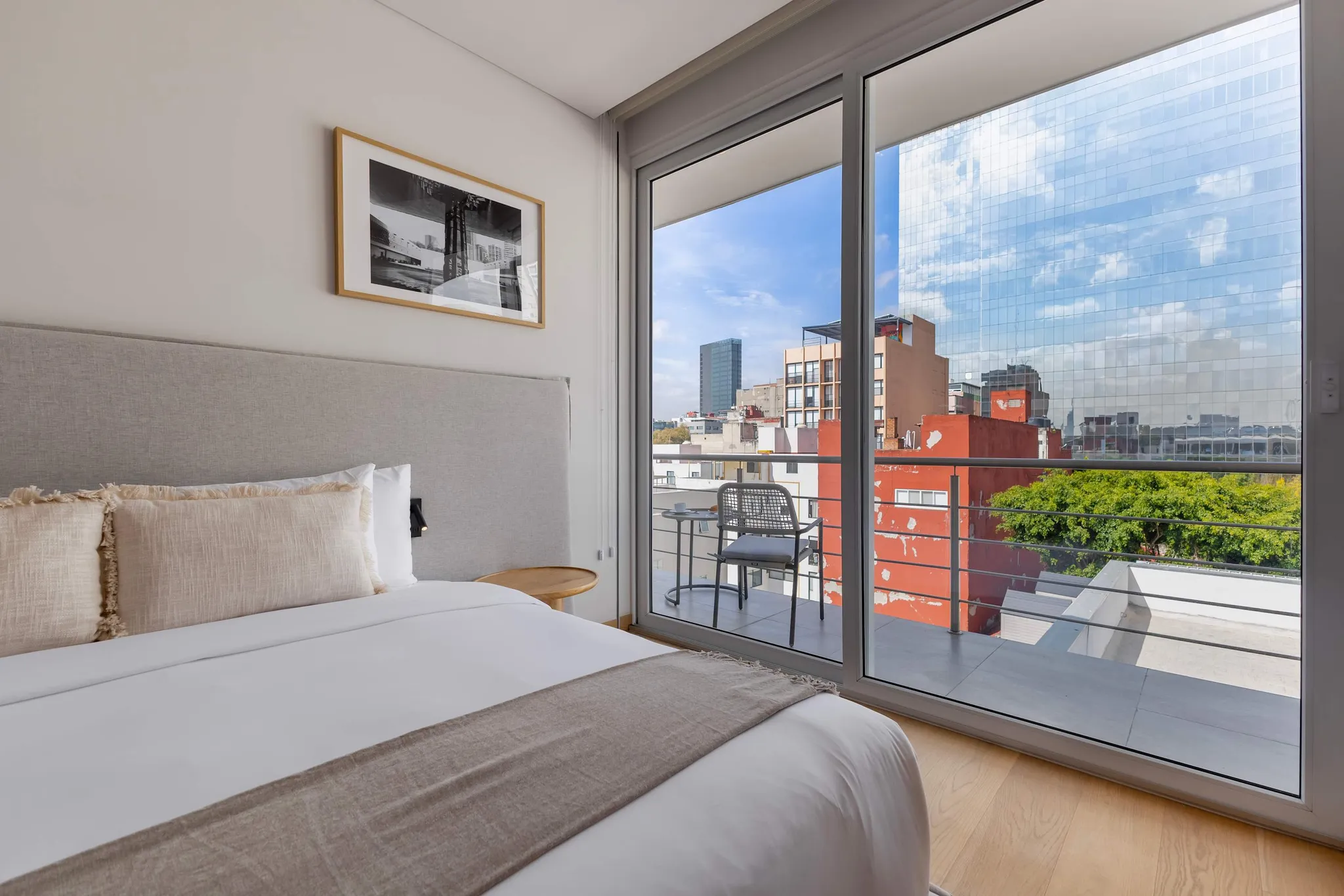 A bed with white linens and a grey throw blanket faces a balcony with chairs and a table.