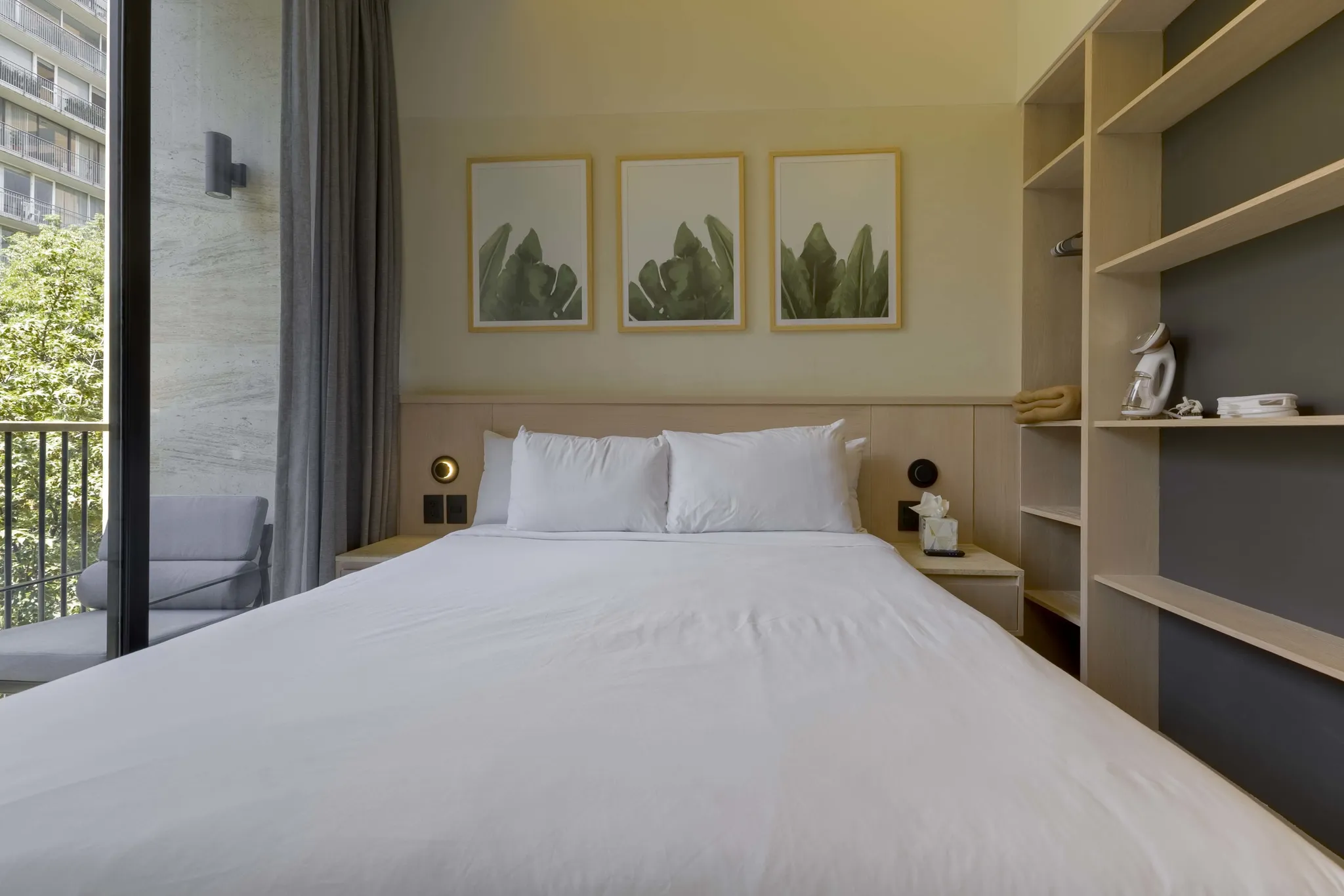 A bed with white linens is framed by a modern shelving unit and natural light.