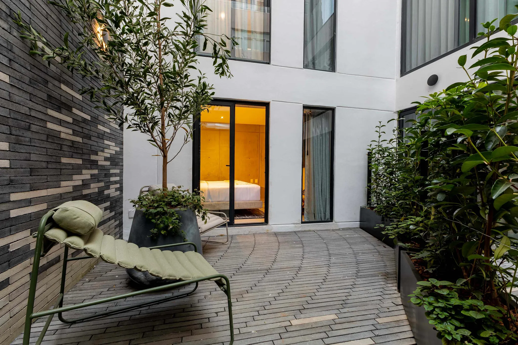 A modern courtyard with a brick wall, chaise lounge, and lush greenery.